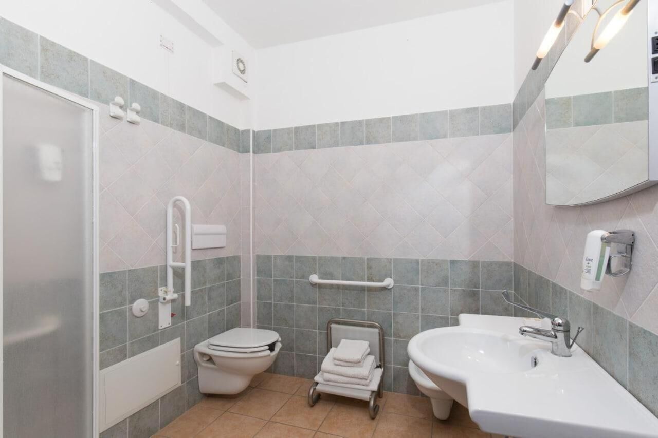 A modern bathroom with a toilet, sink, and shower. It features grab bars, a towel rack, and a mirror.