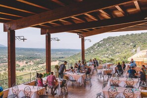 An outdoor dining area with tables and chairs, wooden beams overhead, and a view of a hilly landscape. - Hotel Ispinigoli (Dorgali)