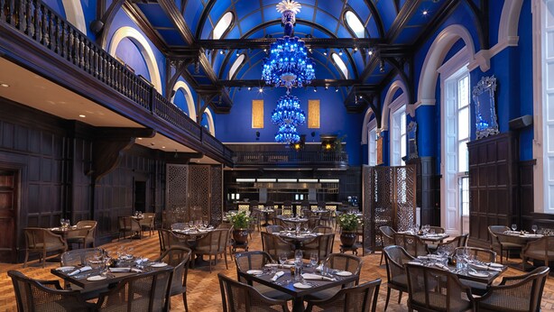 A spacious dining area with multiple tables and chairs, a grand chandelier, and a high ceiling with arched windows. - The Lalit London (London)