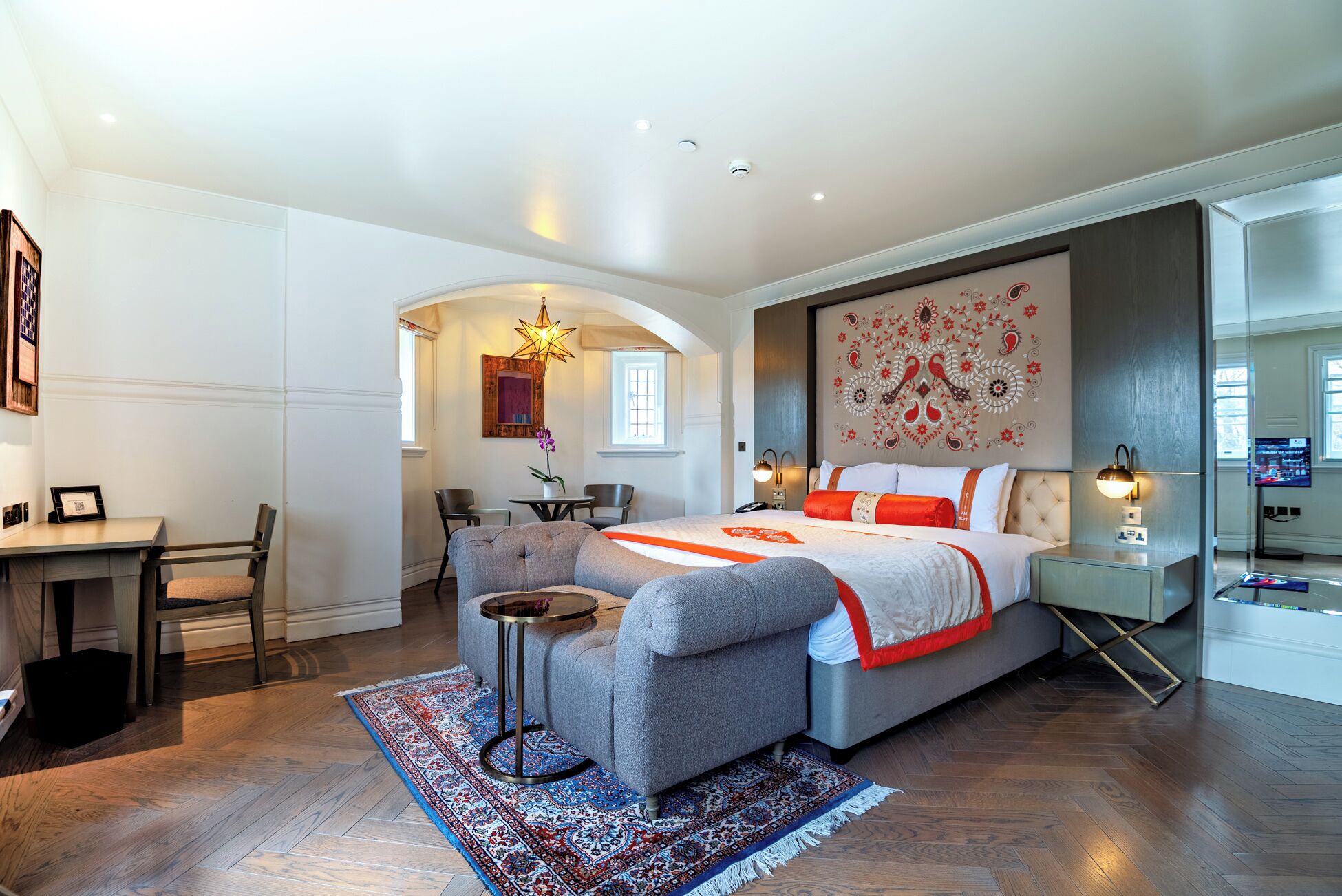 A modern bedroom with a large bed, a desk, and a chair. There is a decorative wall piece and a television on the wall.