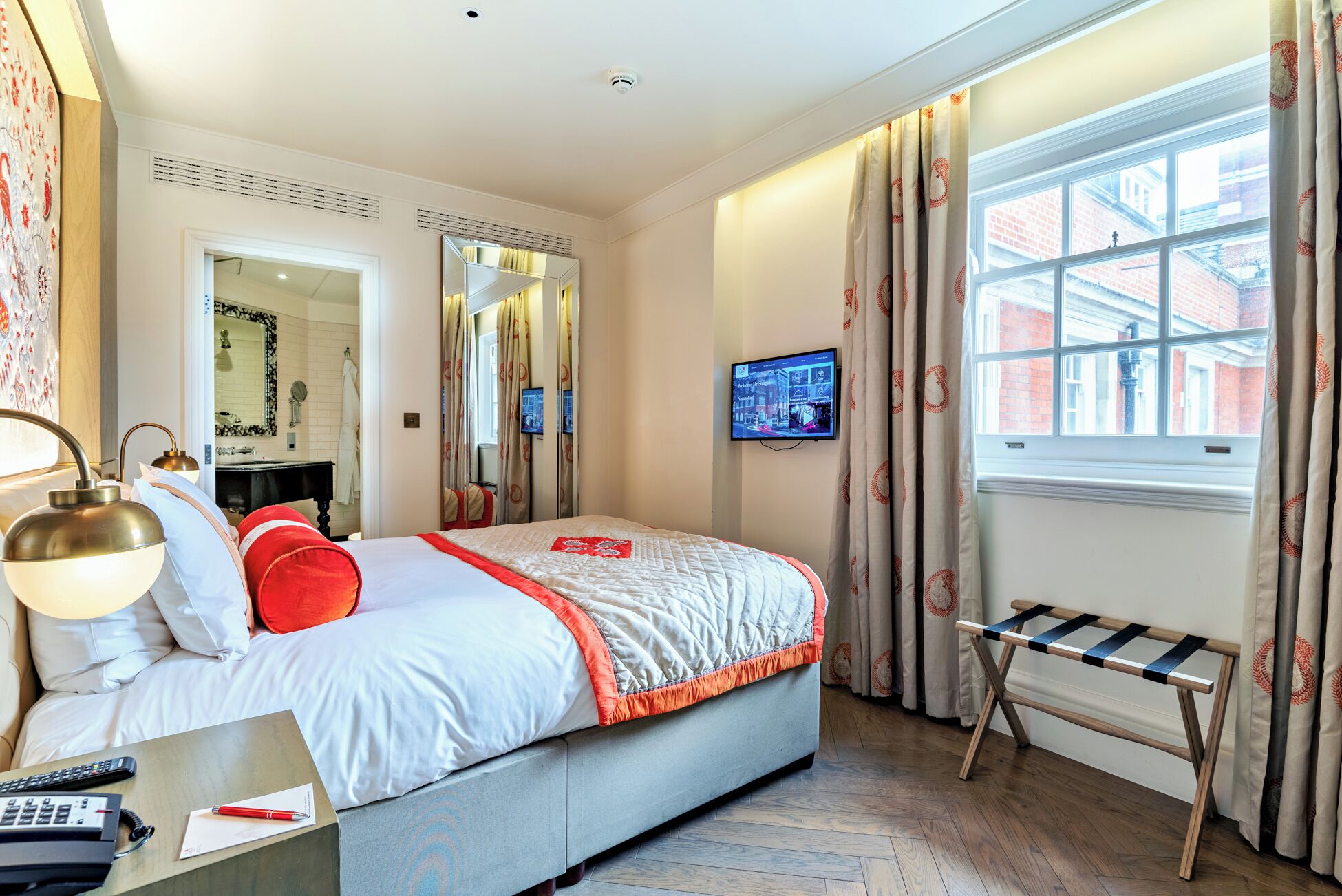 A hotel room with a bed, a bedside table with a lamp, a TV on the wall, and a window with curtains.