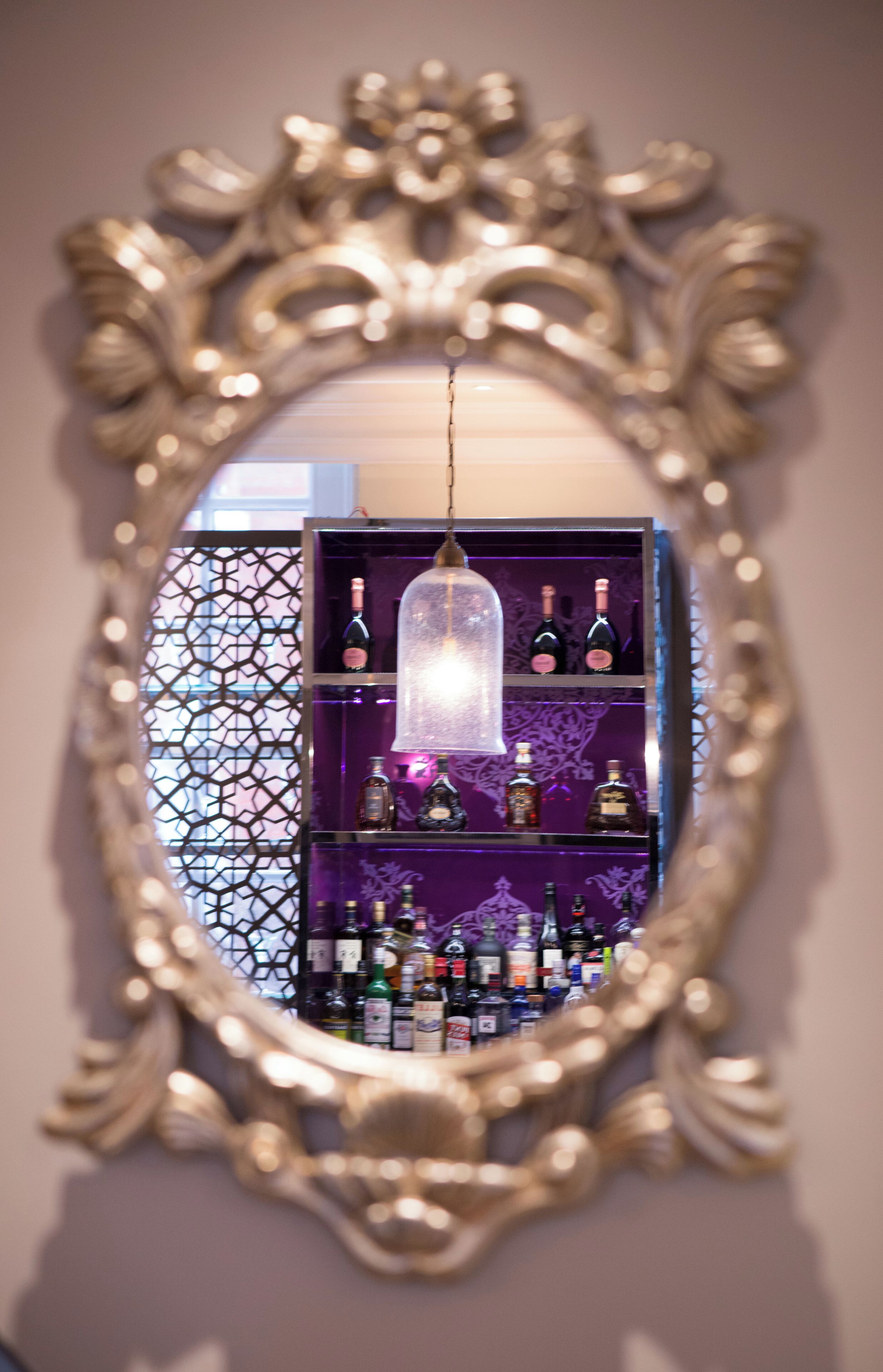 Un miroir doté d’un cadre orné, qui reflète un coin bar avec des étagères remplies de bouteilles.