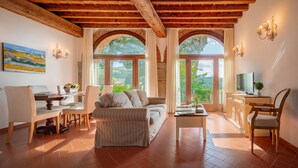 A spacious living area with a wooden ceiling, a sofa, a coffee table, and a TV on a cabinet. There are two windows with curtains, a painting on the wall, and a chair. - Casabella Residenza d'Epoca (Vaglia)