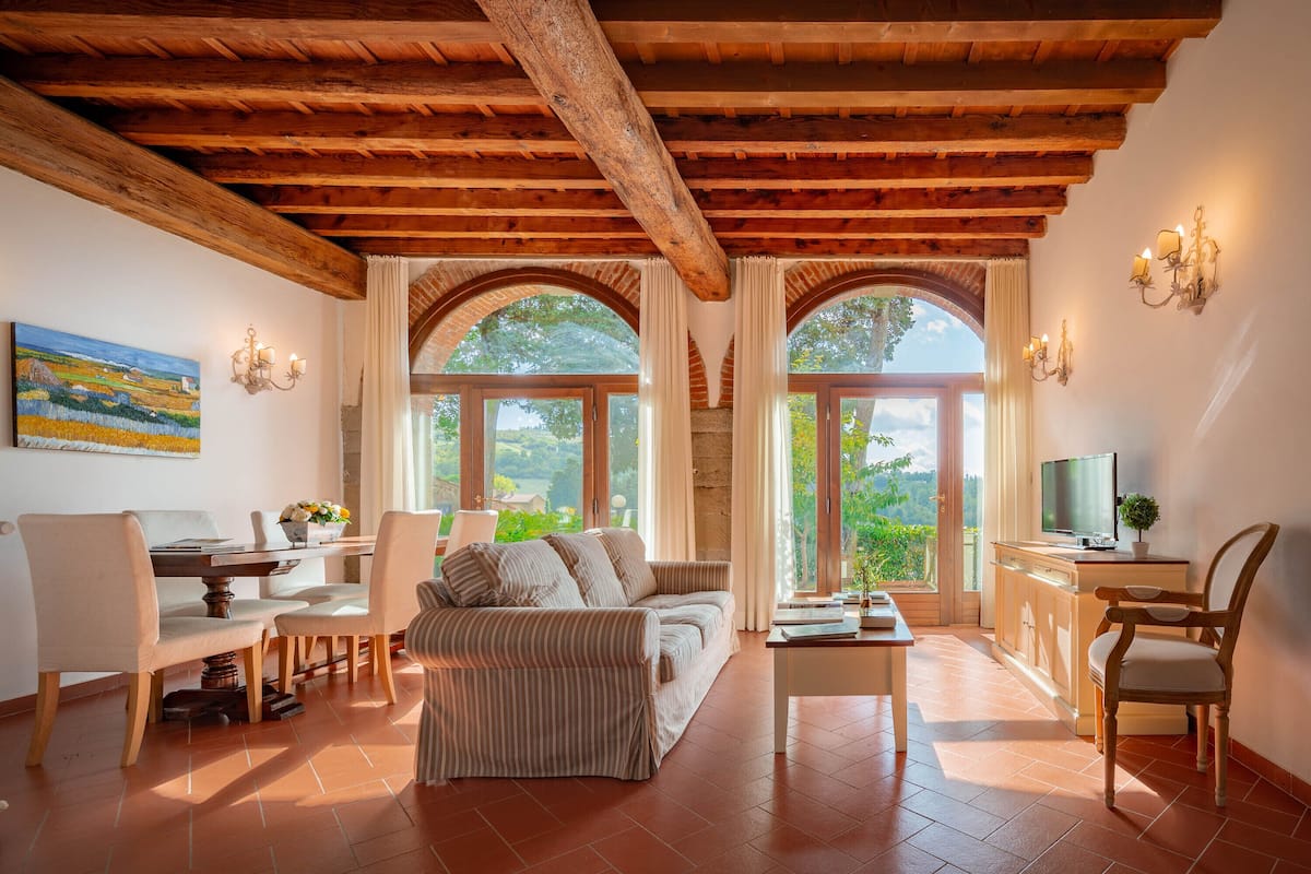 A spacious living area with a wooden ceiling, a sofa, a coffee table, and a TV on a cabinet. There are two windows with curtains, a painting on the wall, and a chair.