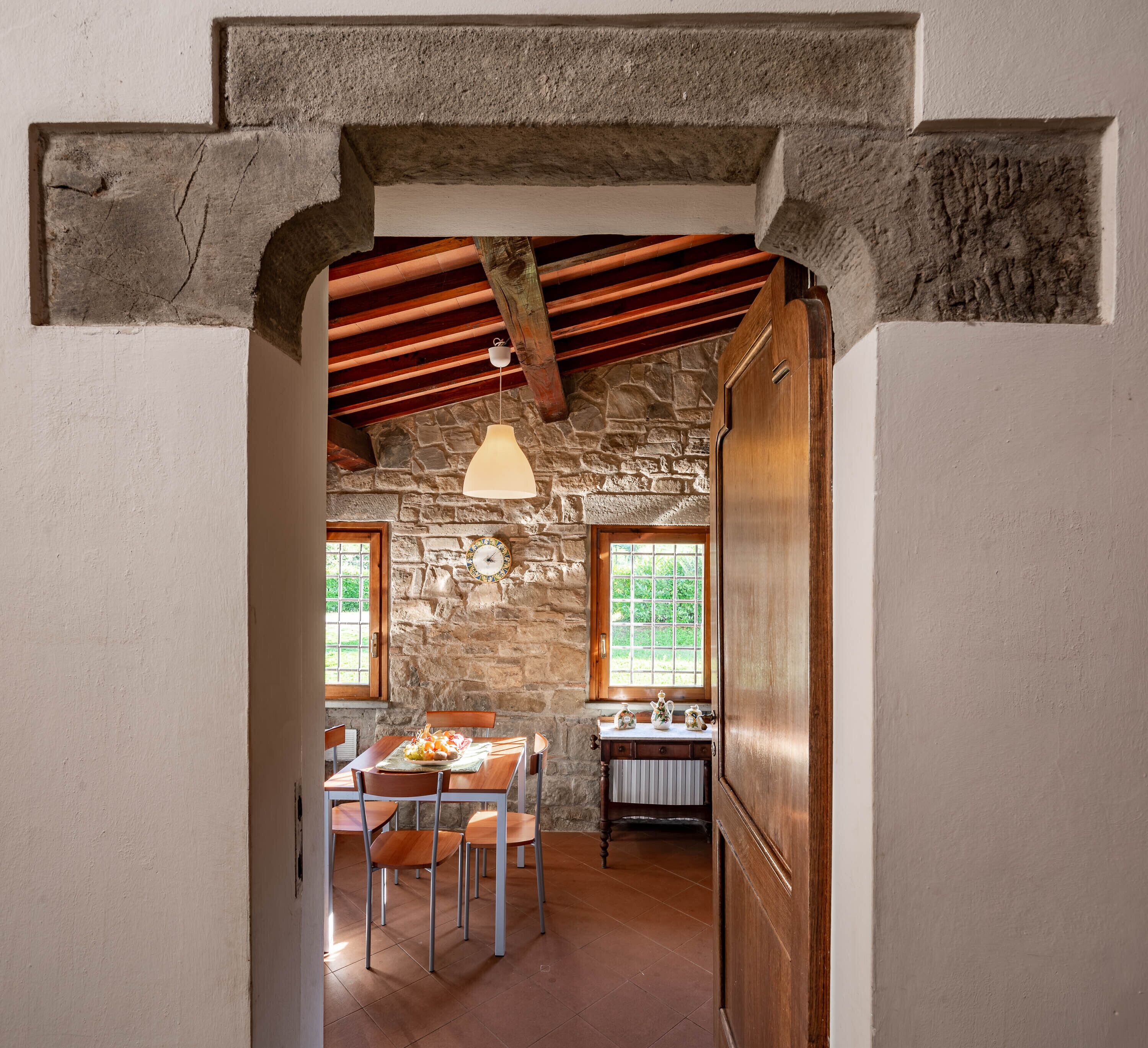 Una habitación rústica con paredes de piedra, una mesa y sillas de madera, una lámpara colgante y una ventana con vistas a la vegetación.