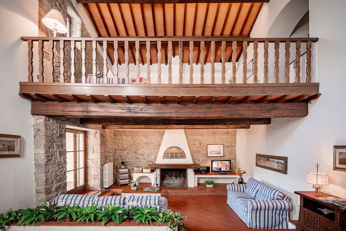A spacious living area with a high ceiling, exposed wooden beams, a stone fireplace, and a comfortable seating area with striped cushions.
