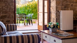 A cozy room with a striped sofa, a wooden coffee table with a book and a flower vase, and a view of an outdoor patio with chairs and greenery. - Casabella Residenza d'Epoca (Vaglia)