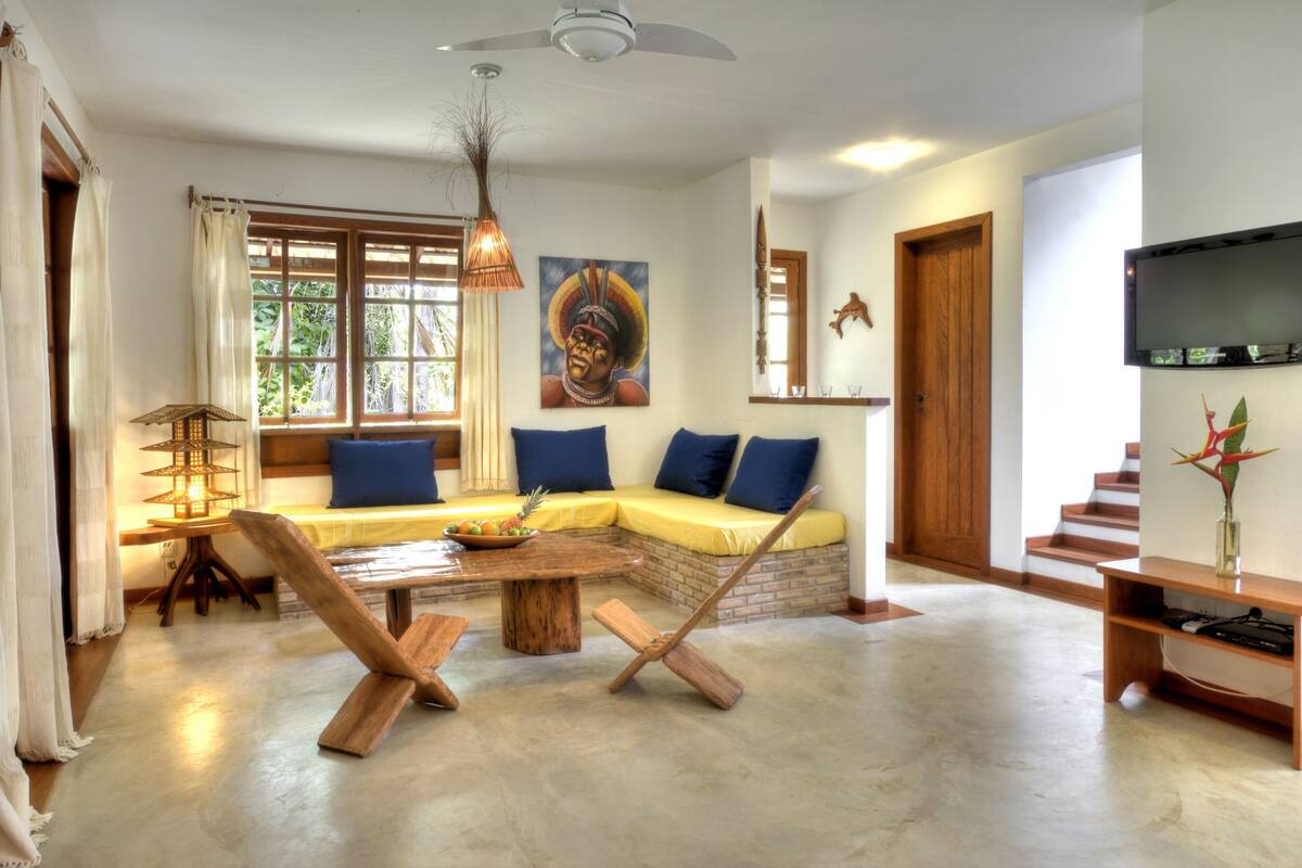 A living room with a wooden bench, a sofa with blue cushions, a dining table, and a TV on the wall.