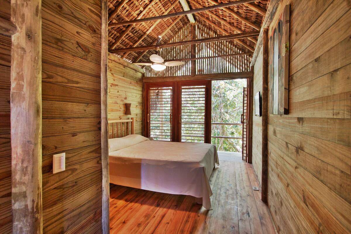 A wooden cabin bedroom with a bed, a ceiling fan, and a view of the outdoors through a door.