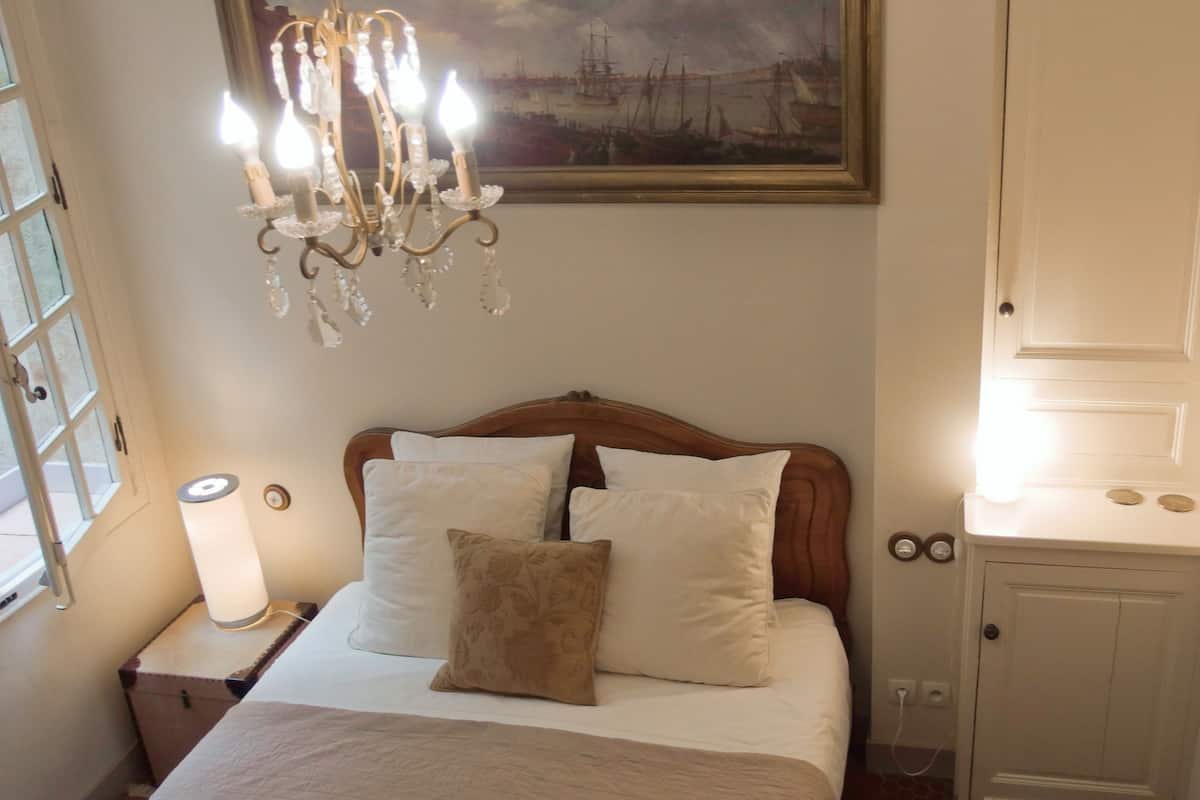 A bedroom with a wooden bed, white bedding, a chandelier, a painting, and a small bedside table with a lamp.