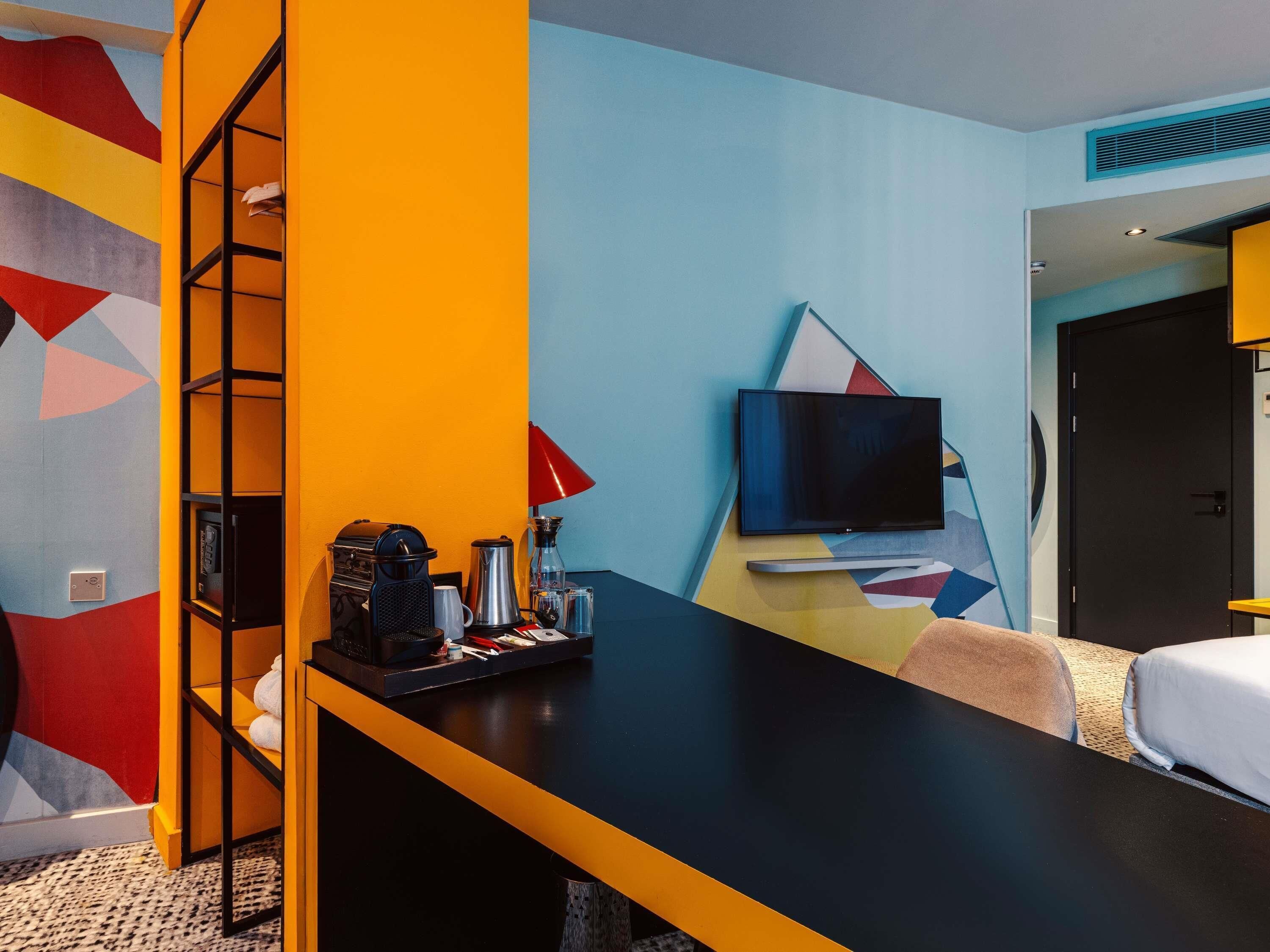 A hotel room with a bed, a desk with a coffee maker, a TV, and a colorful wall.