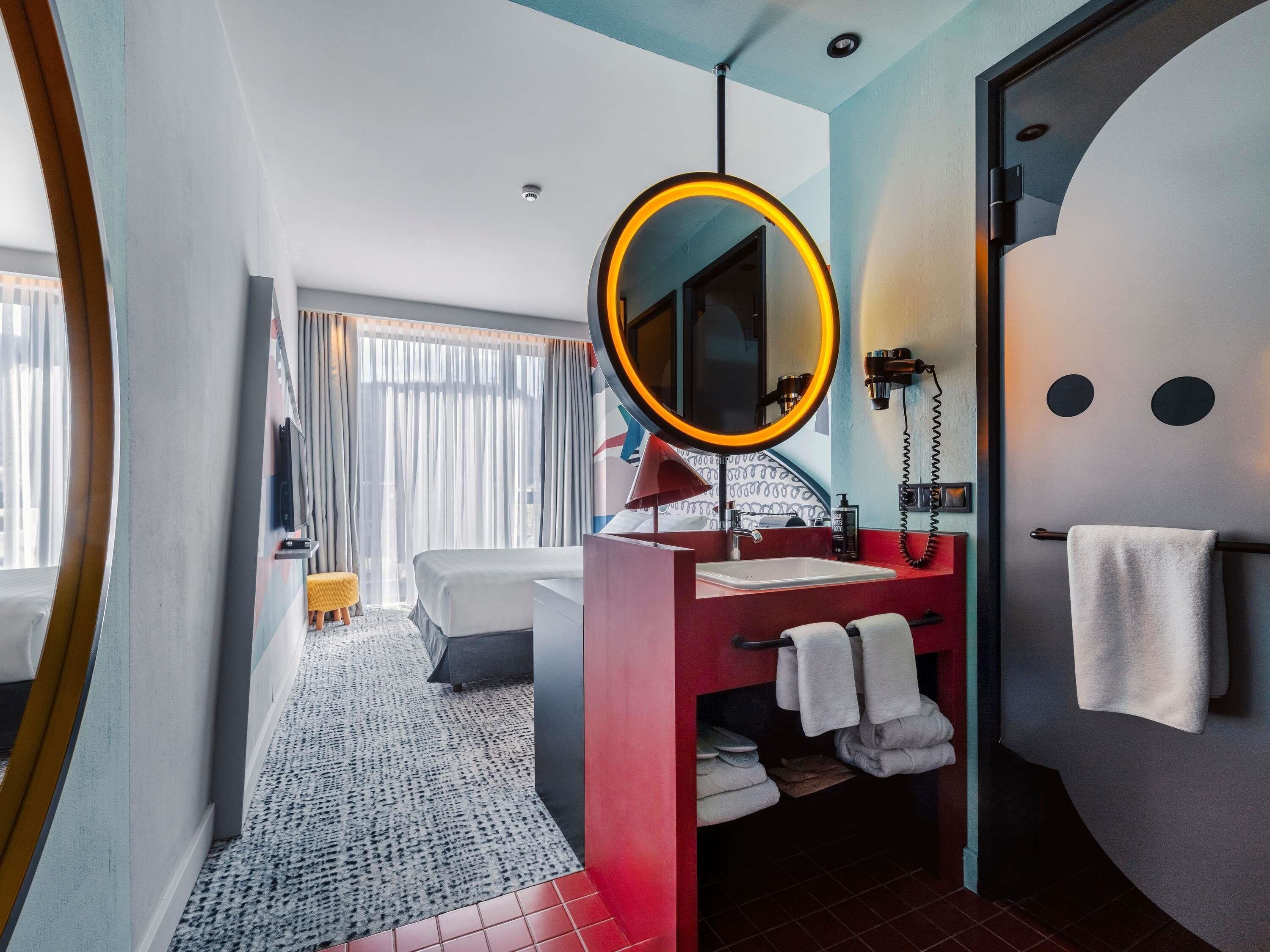 Une chambre d’hôtel avec un grand miroir, une coiffeuse rouge, un lavabo blanc, un porte-serviettes, un lit et une fenêtre avec des rideaux.