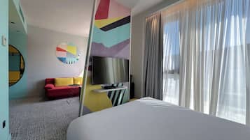 A hotel room with a bed, a TV, a red sofa, a small table, and a colorful abstract wall art.