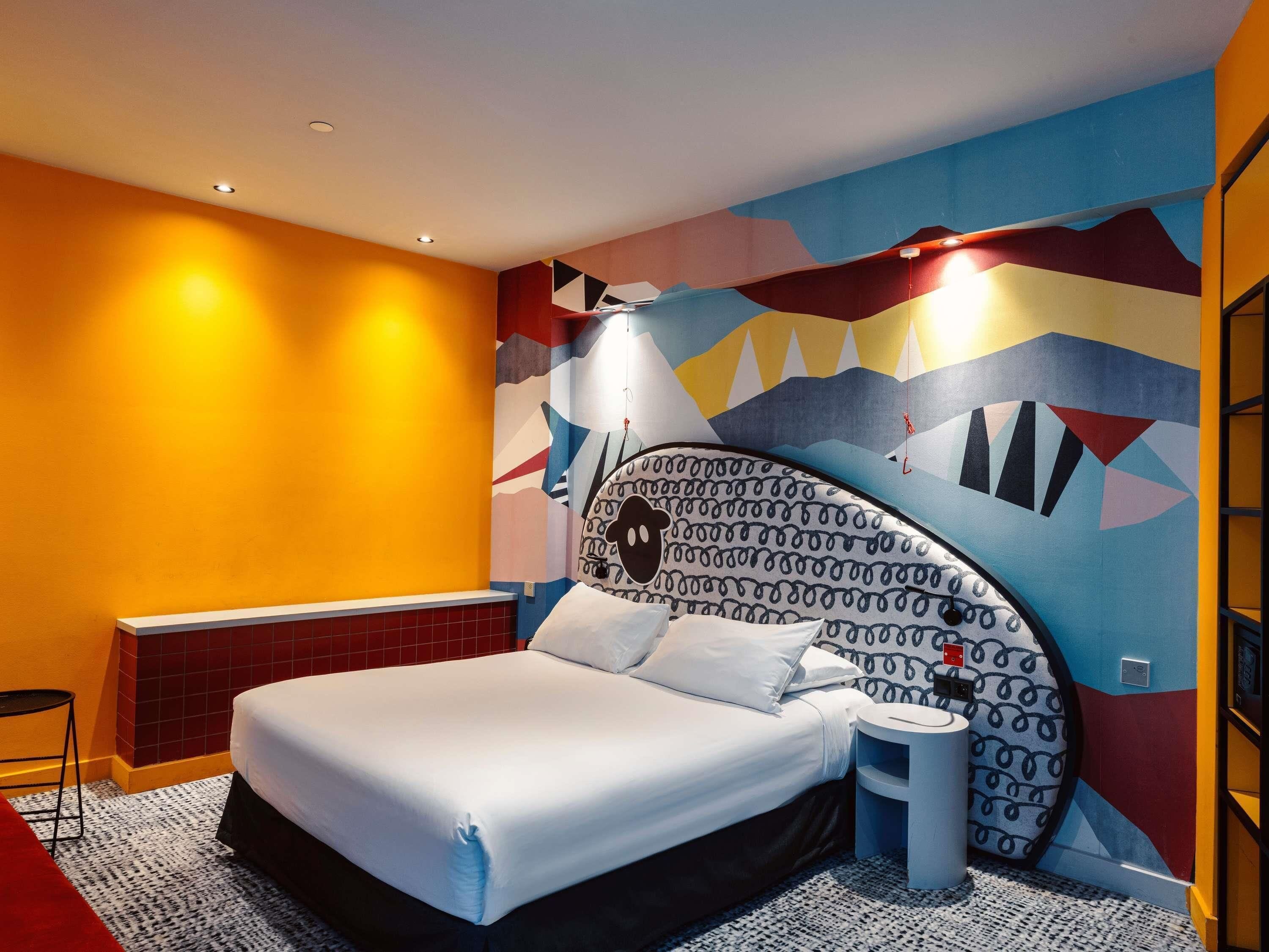 Ein modernes Hotelzimmer mit einem großen Bett, einem kleinen Nachttisch und einem farbenfrohen, abstrakten Wandbild.