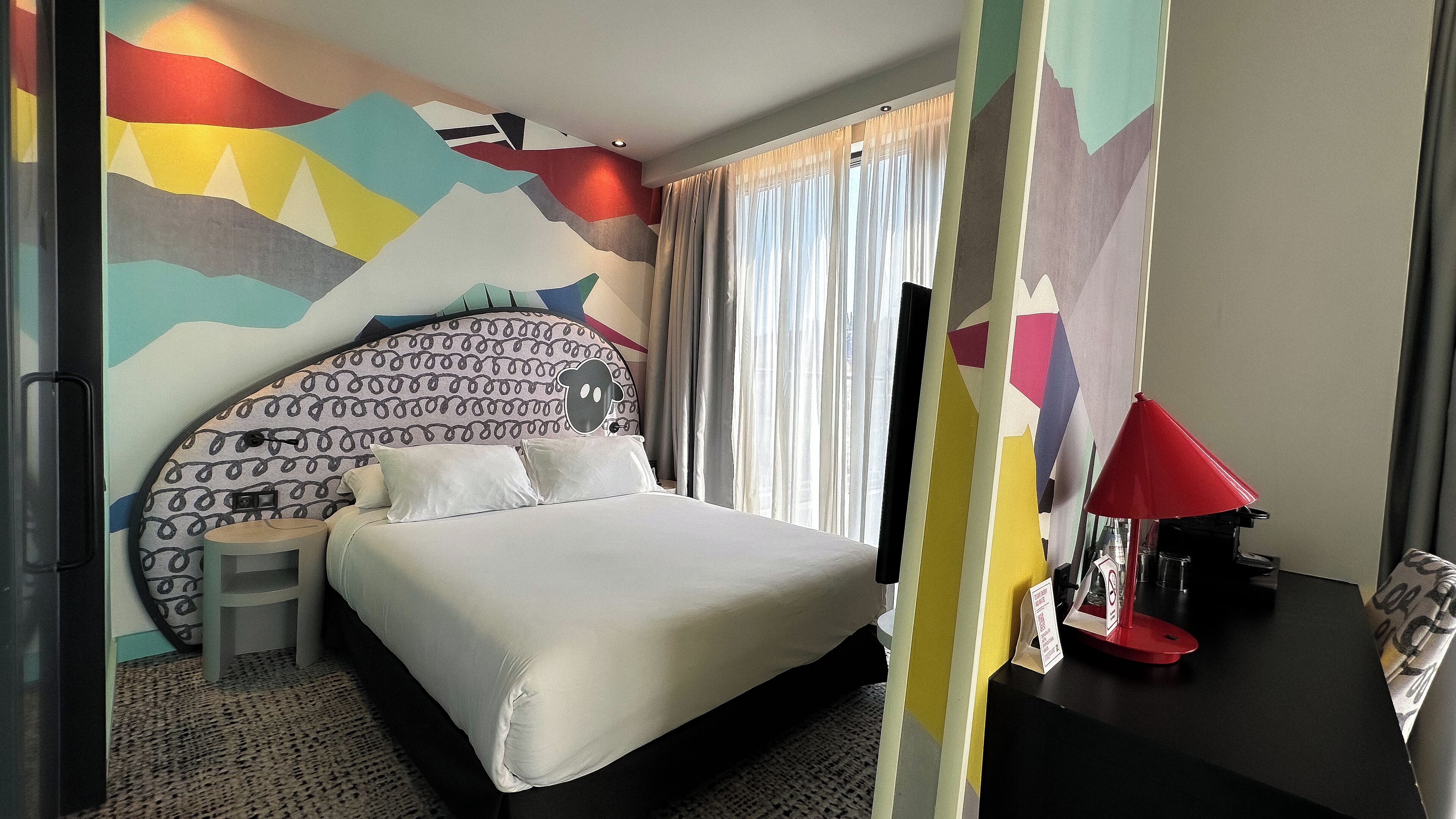 A hotel room with a bed, a nightstand, a lamp, and a wall with a colorful abstract design.