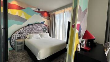 A hotel room with a bed, a nightstand, a lamp, and a wall with a colorful abstract design.