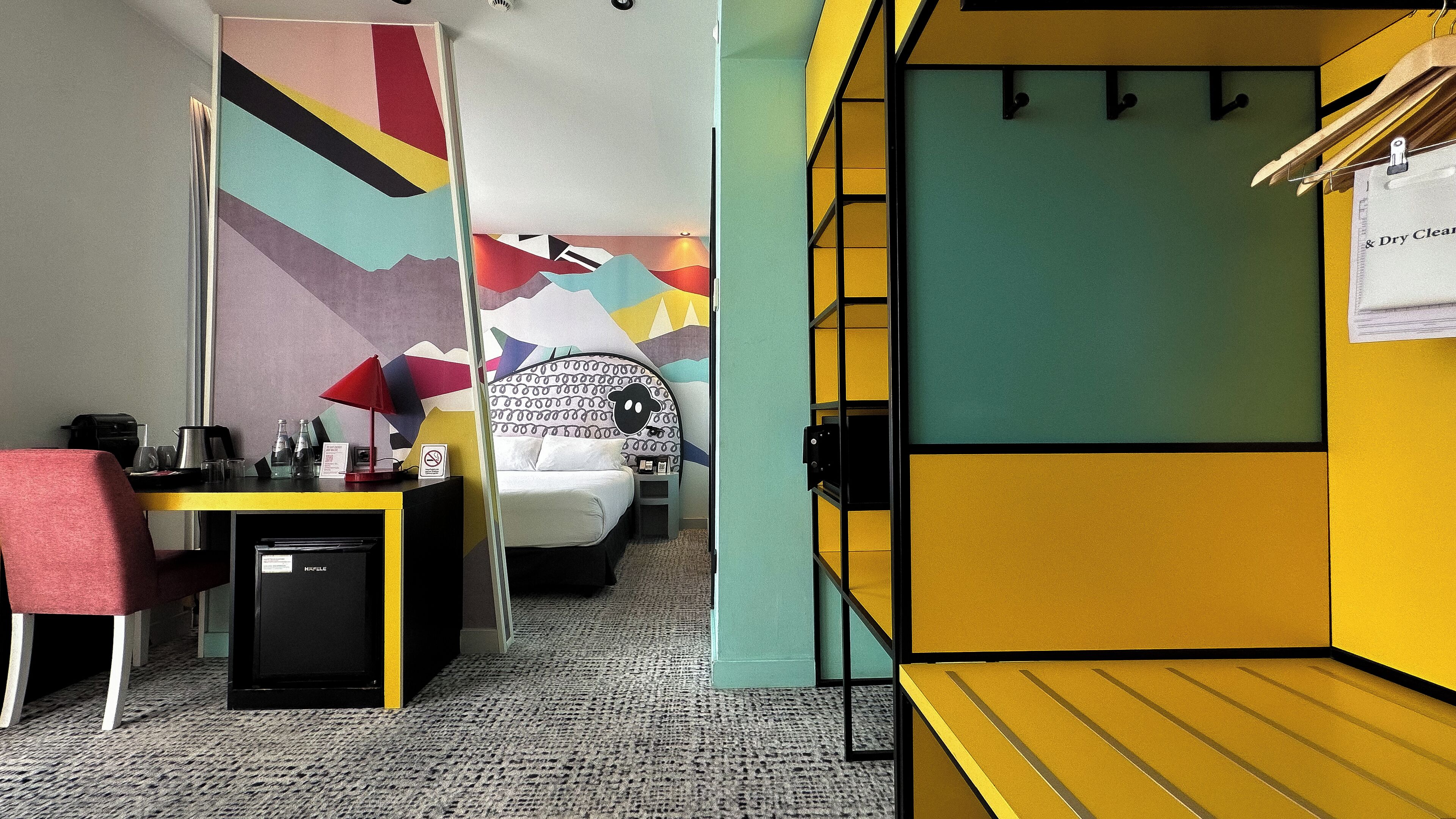 Hotellihuone, jossa on sänky, työpöytä tuolilla, kahvinkeitin ja seinä, jossa on värikäs, geometrinen kuvio.