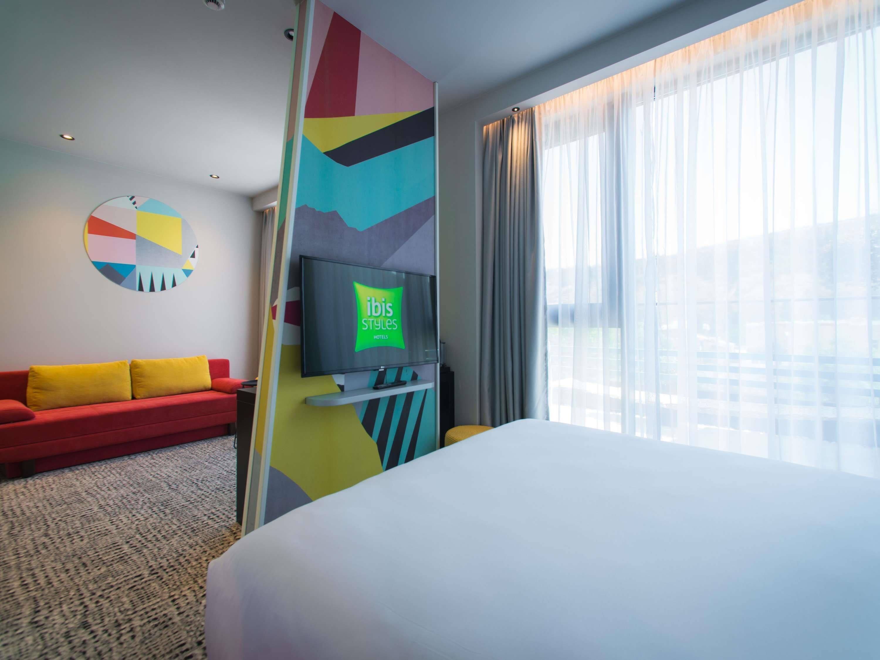 Ein Hotelzimmer mit Bett, Fernseher, einem roten Sofa, farbenfroher Wandkunst und einem Fenster mit Vorhängen.