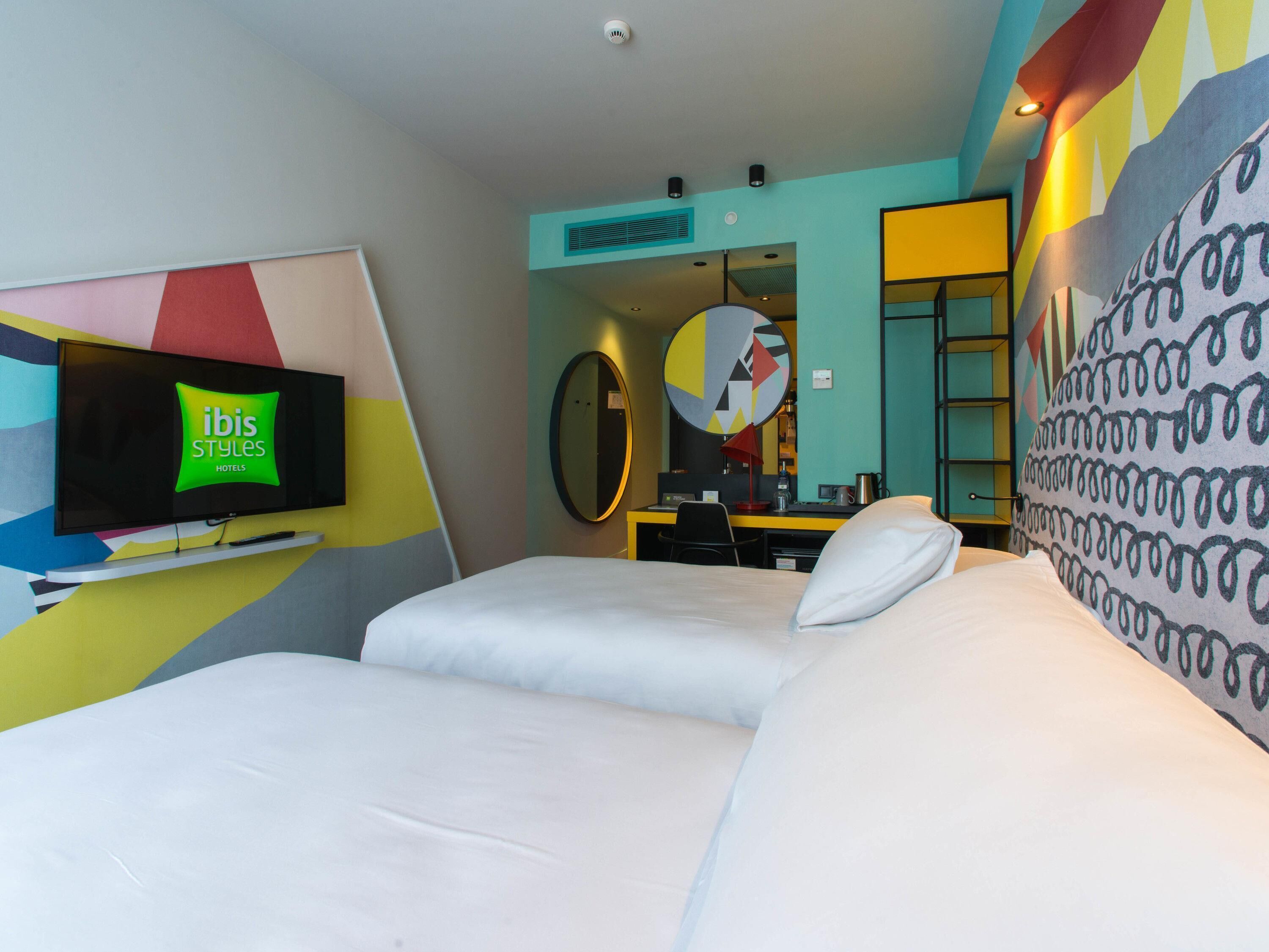 A hotel room with a bed, a desk, a chair, a TV mounted on the wall, and a mirror.