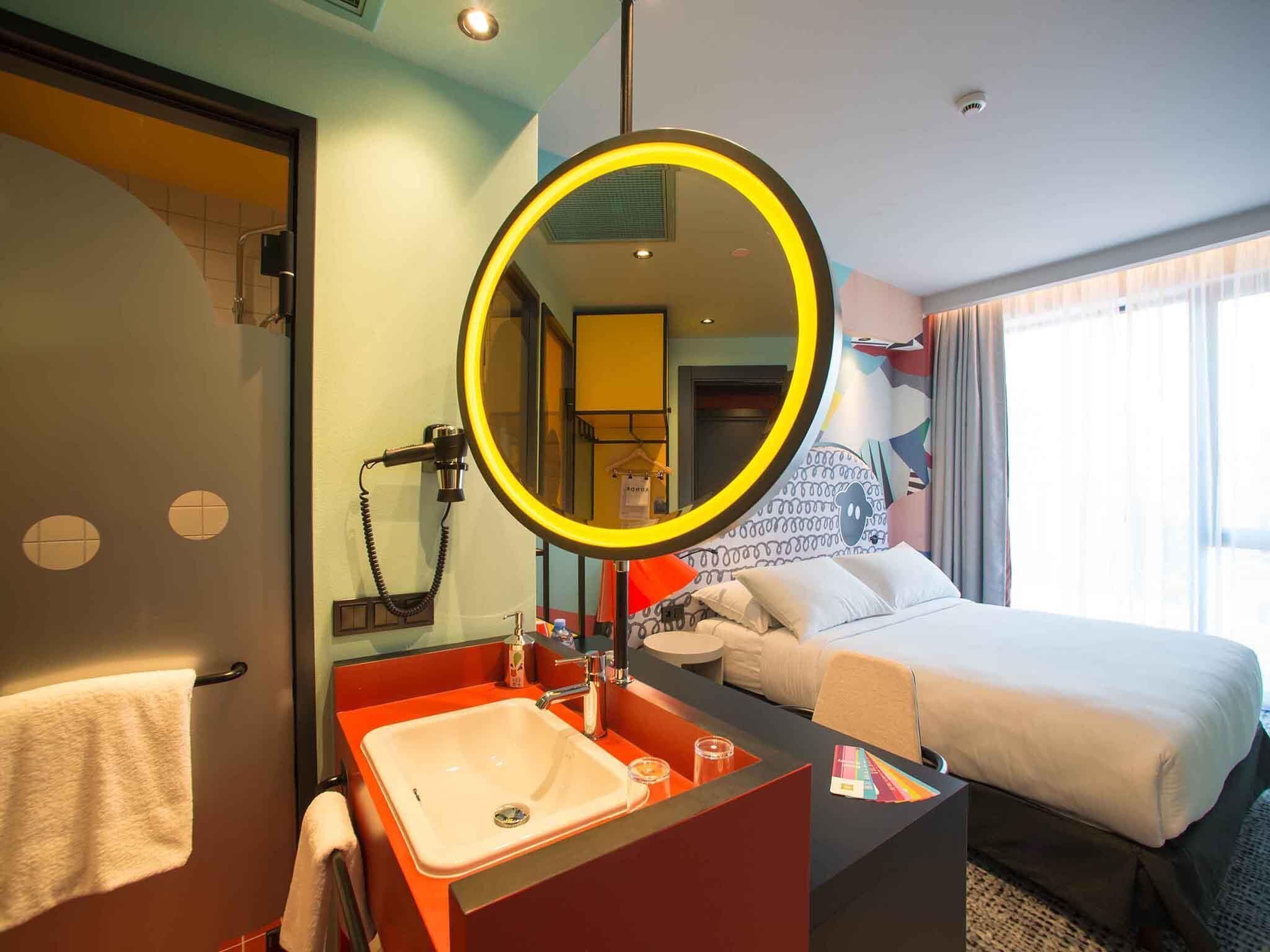 Une chambre d’hôtel avec un lit, un miroir rond, un lavabo et une fenêtre avec des rideaux.