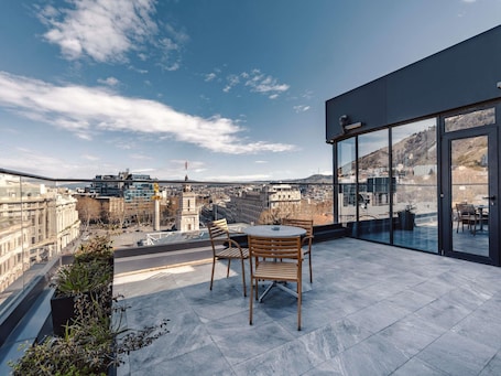 A rooftop terrace with a table and chairs, overlooking a cityscape with buildings and a mountain.