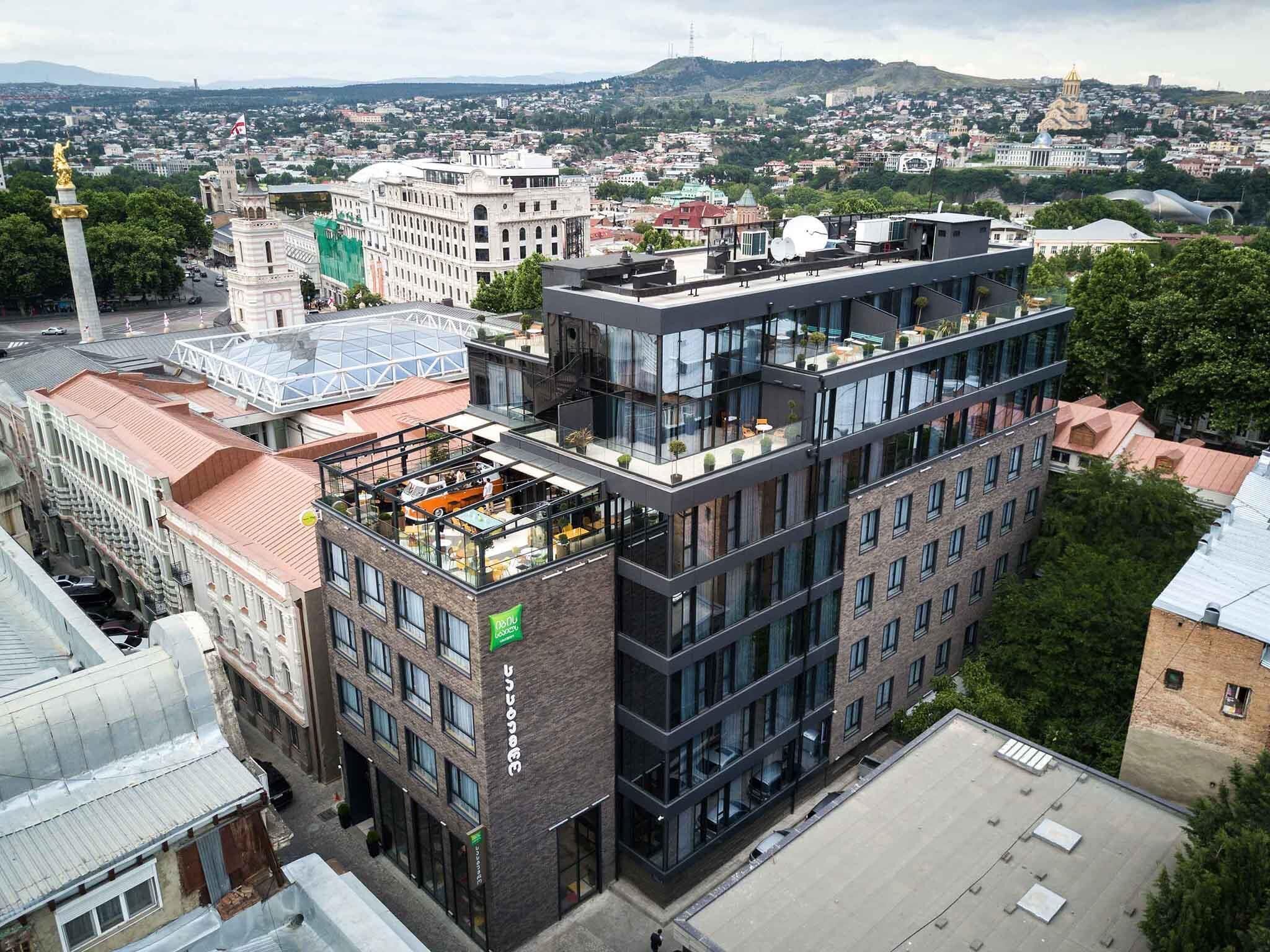 Un edificio de varios pisos con terraza en la azotea, rodeado de otros edificios y un paisaje urbano al fondo.