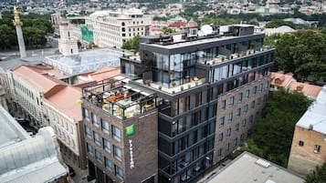 Un edificio de varios pisos con terraza en la azotea, rodeado de otros edificios y un paisaje urbano al fondo.