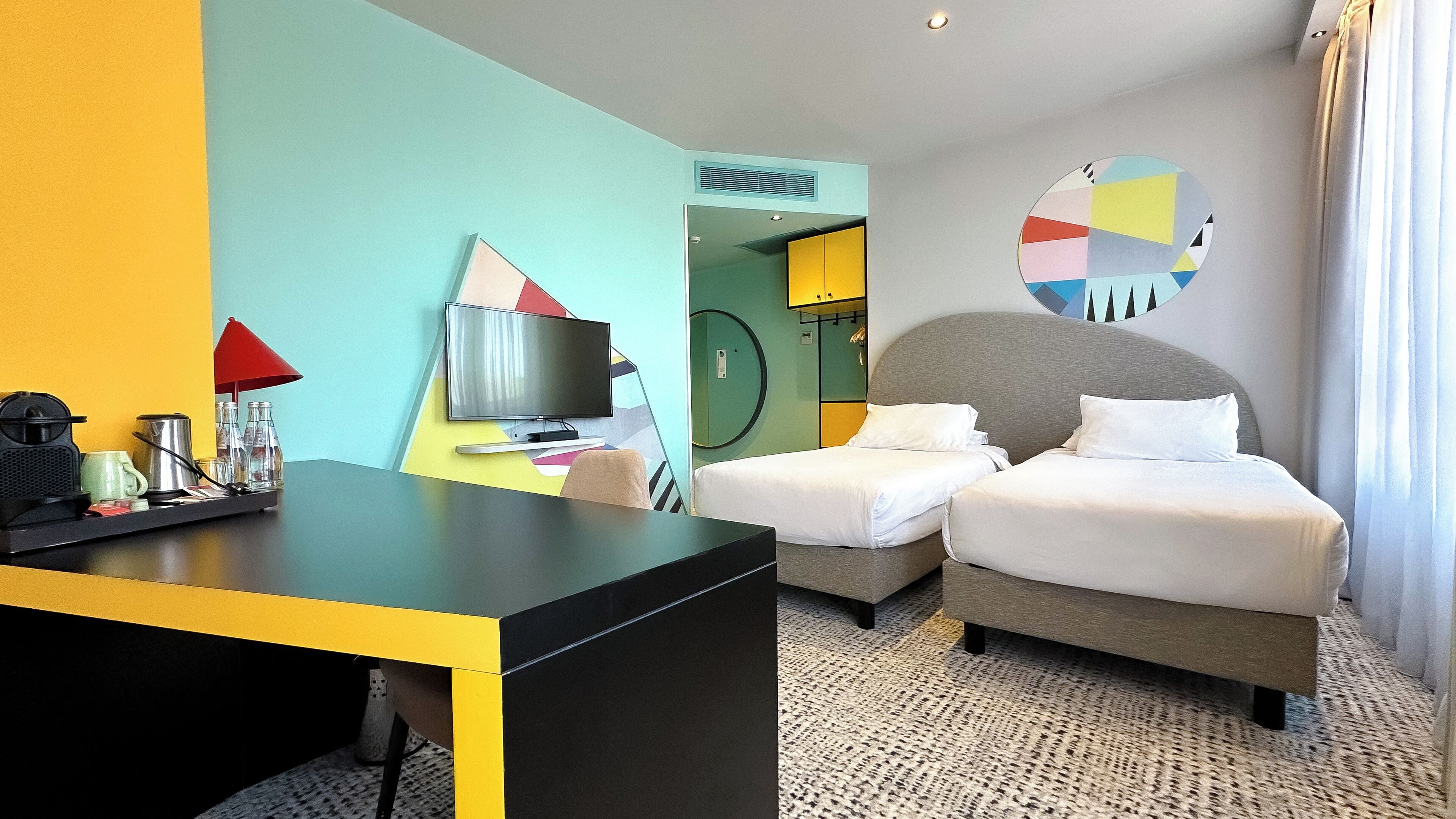 A hotel room with a desk, two beds, a TV, and a circular wall art piece.