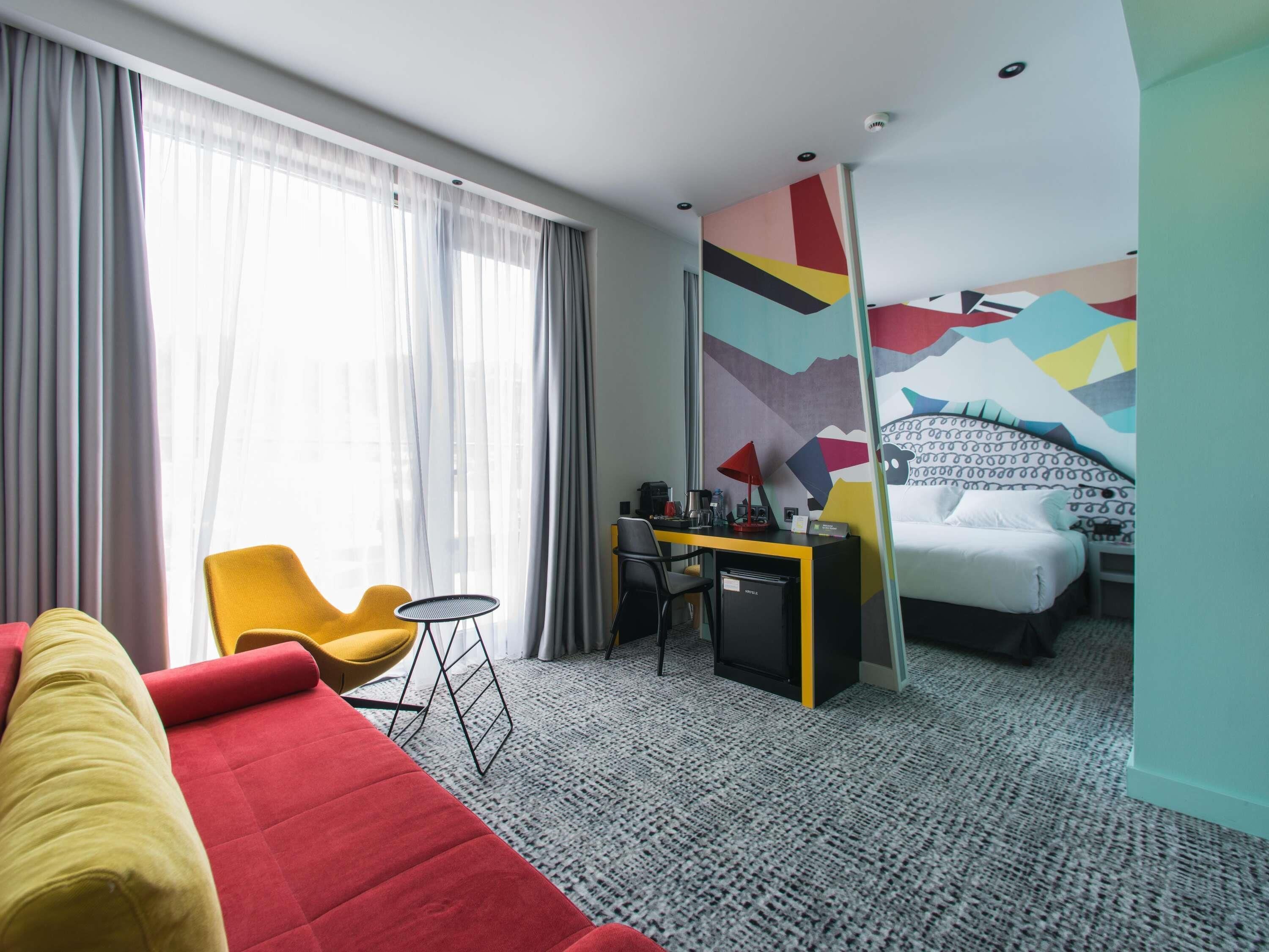 A hotel room with a bed, a desk, a yellow chair, a red sofa, and a colorful wall mural.