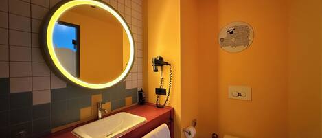 A bathroom with a round mirror, a sink, a hairdryer, a toilet, and a towel rack.