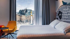 A hotel room with a bed, a chair, a view of buildings, and a patterned headboard. - Ibis Styles Tbilisi Center (Tbilisi)