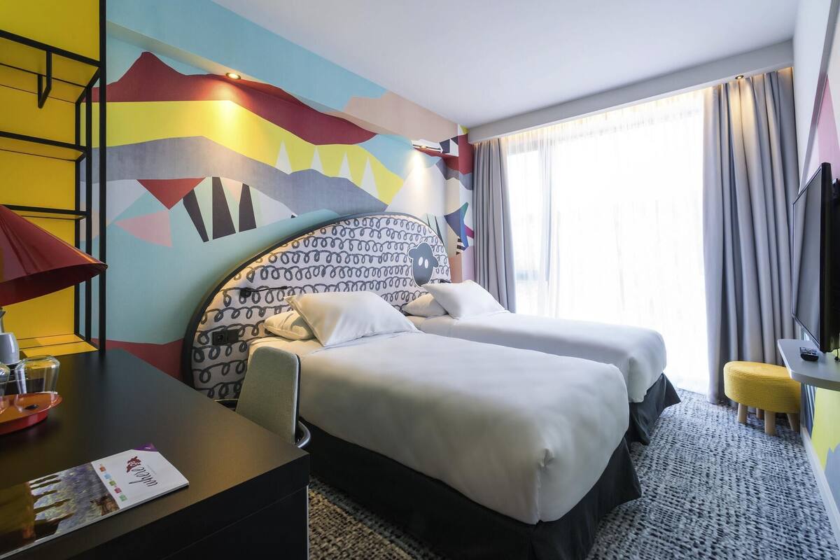 A hotel room with two beds, a desk with a lamp, a TV, and a large window with curtains.