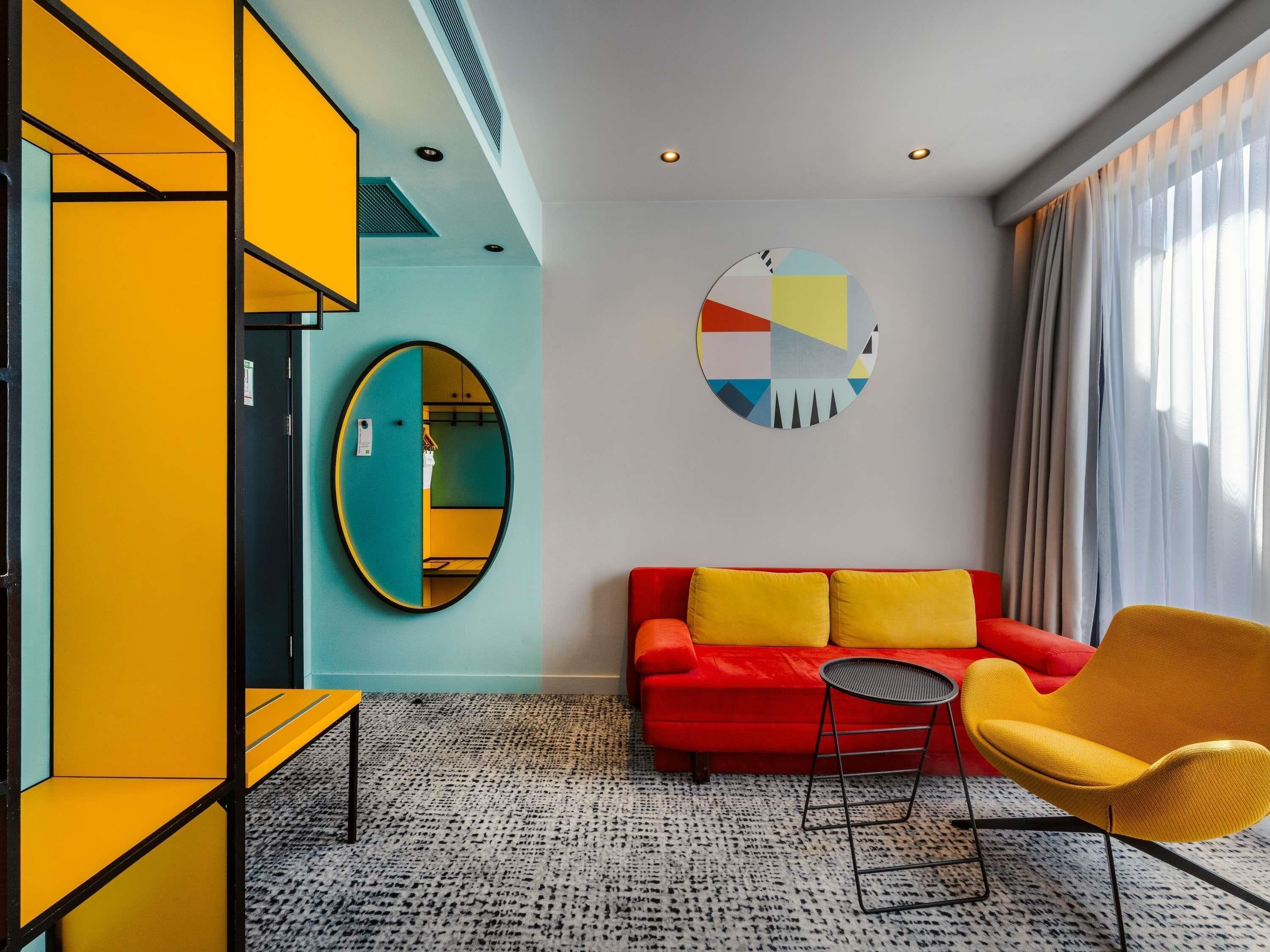 A modern living room with a red sofa, yellow armchairs, a round mirror, and a geometric wall art piece.