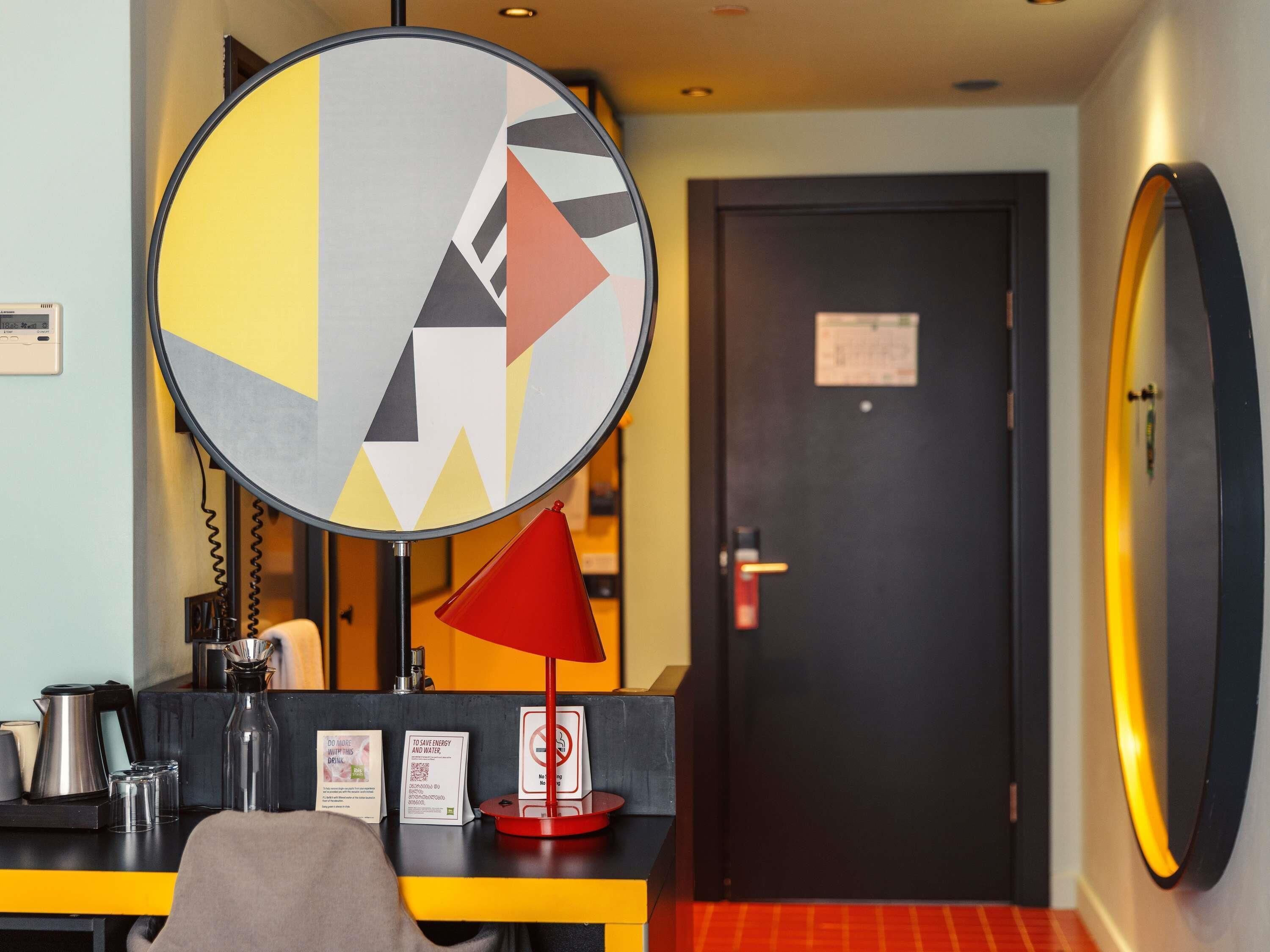 A hotel room with a round mirror, a red lamp, a coffee machine, and a door with a sign.