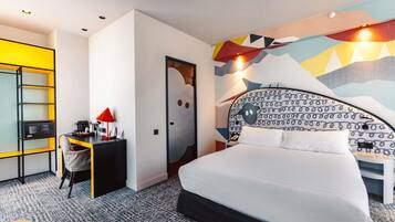 A hotel room with a bed, a desk with a computer, a chair, and a small table with a lamp.