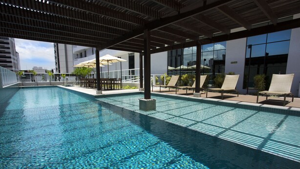 Ein Dachpool mit klarem, blauem Wasser, umgeben von Liegestühlen und Sonnenschirmen, mit modernen Gebäuden im Hintergrund.