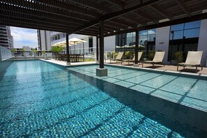 A rooftop swimming pool with clear blue water, surrounded by lounge chairs and umbrellas, and modern buildings in the background.