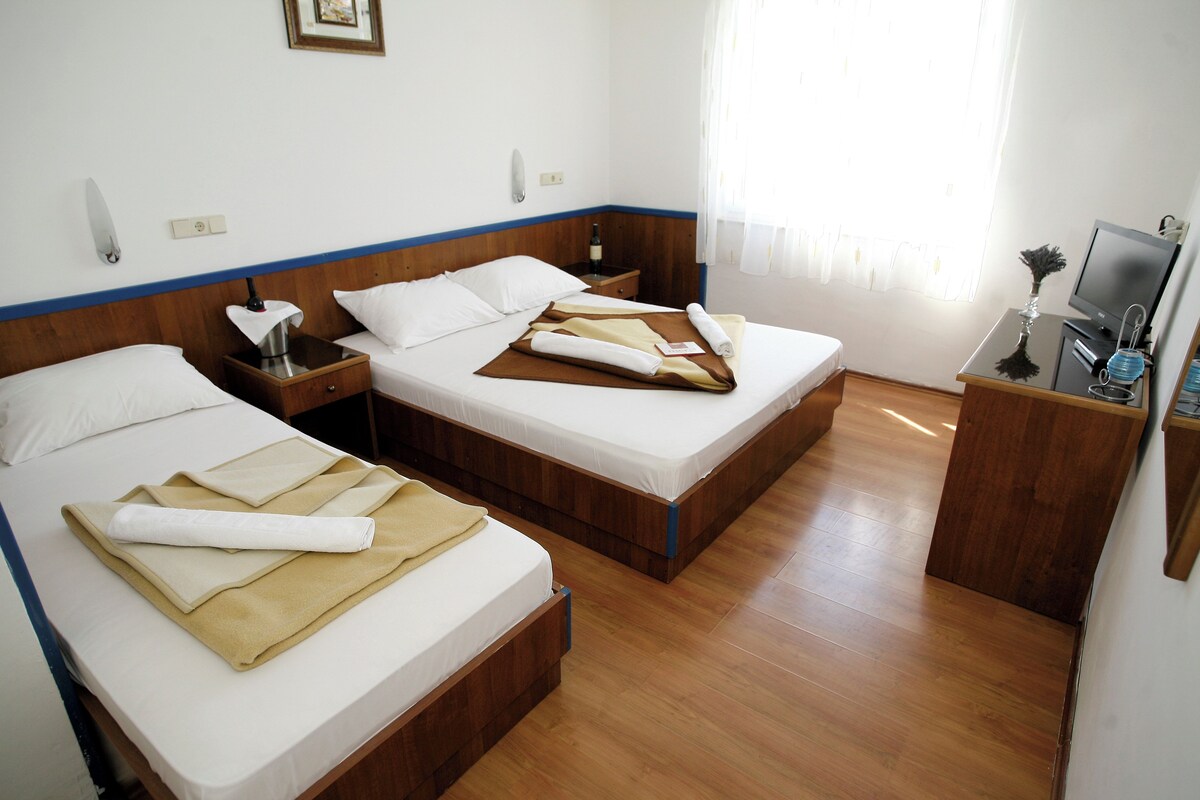 Ein Hotelzimmer mit zwei Betten, Holz-Kopfteilen, einem kleinen Nachttisch mit Lampe, einem Fernseher und einem Fenster mit Vorhängen.