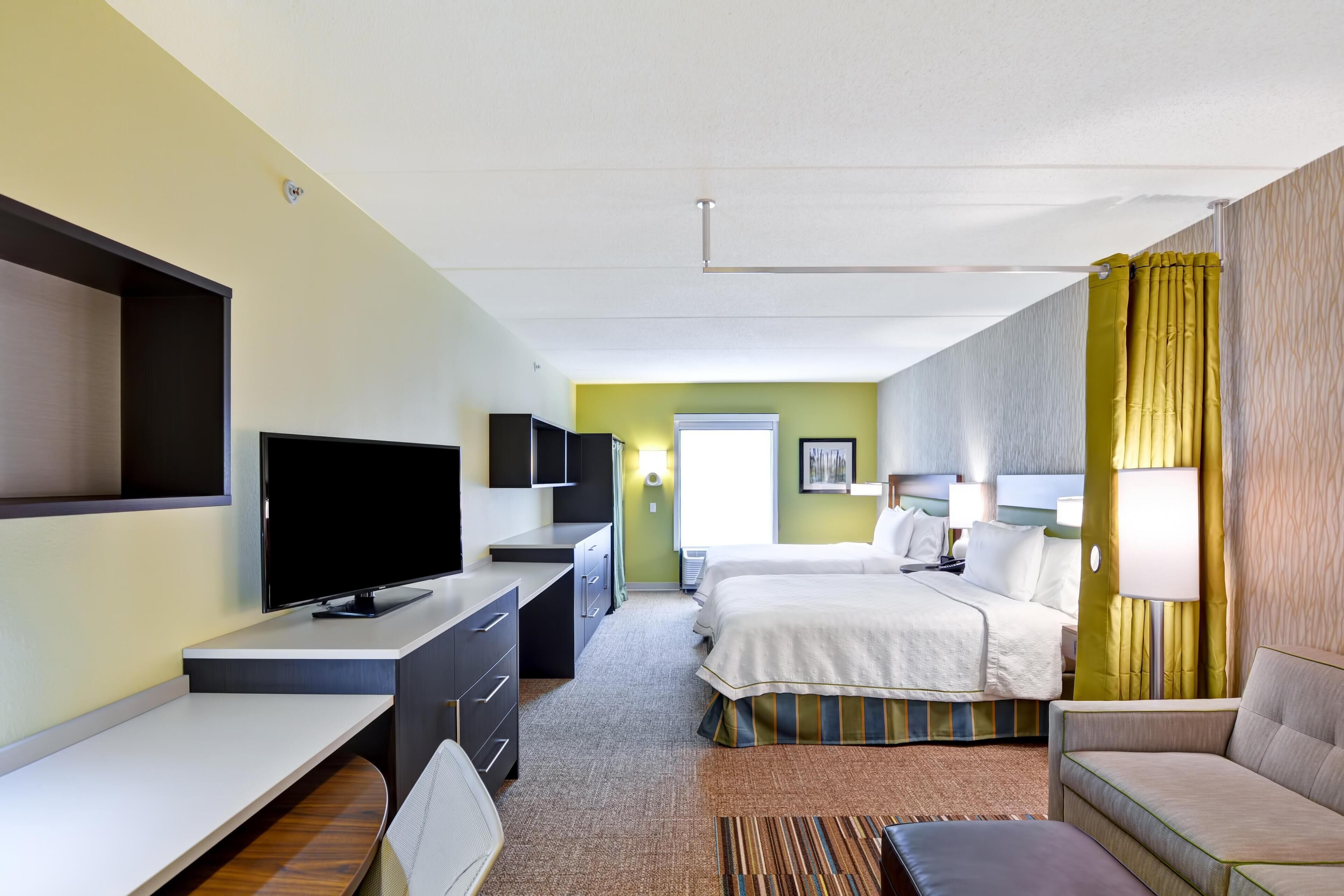 A hotel room with a TV, desk, two beds, a sofa, and a window.