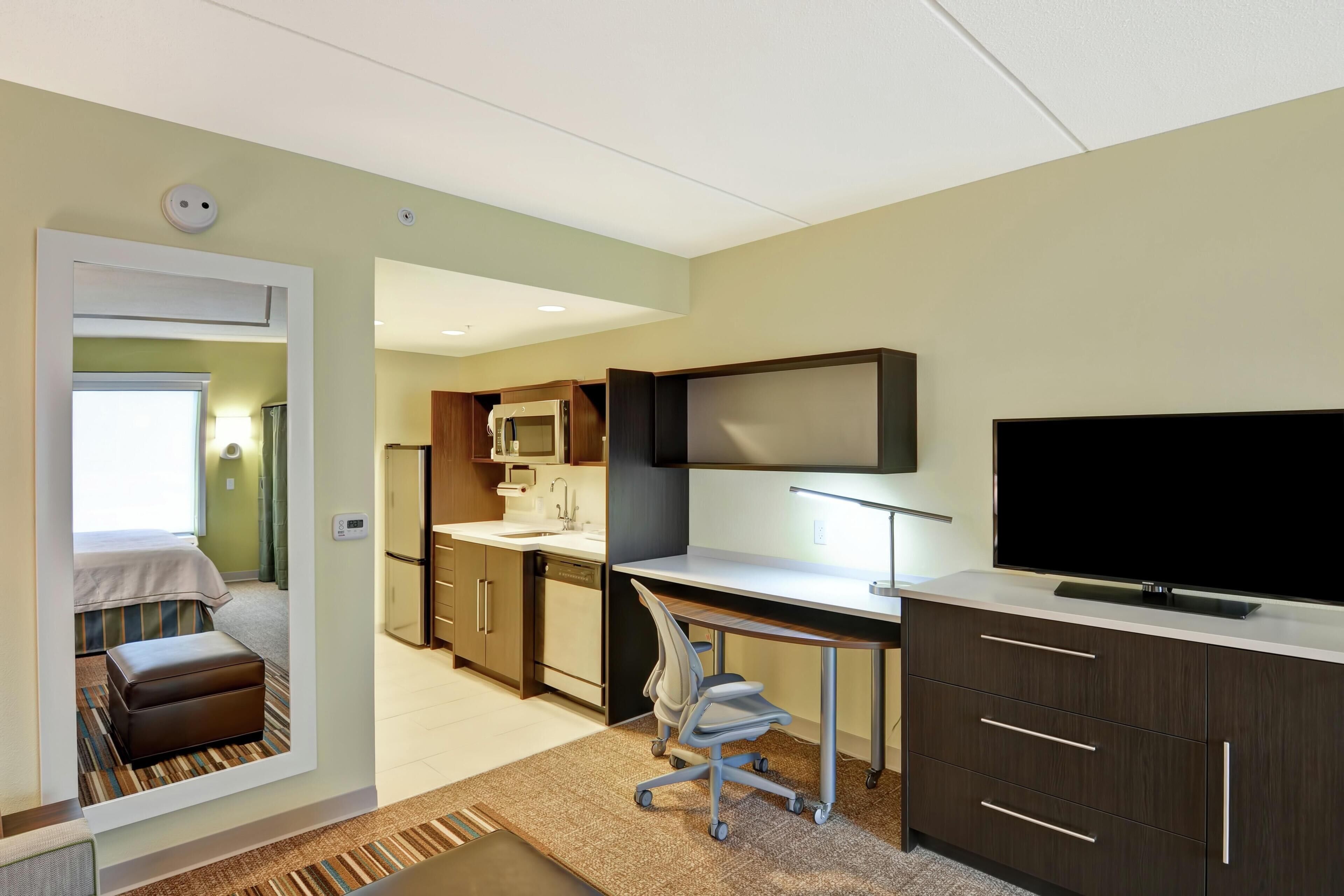 A hotel room with a kitchenette, a bed, a desk with a TV, and a bathroom visible through an open door.