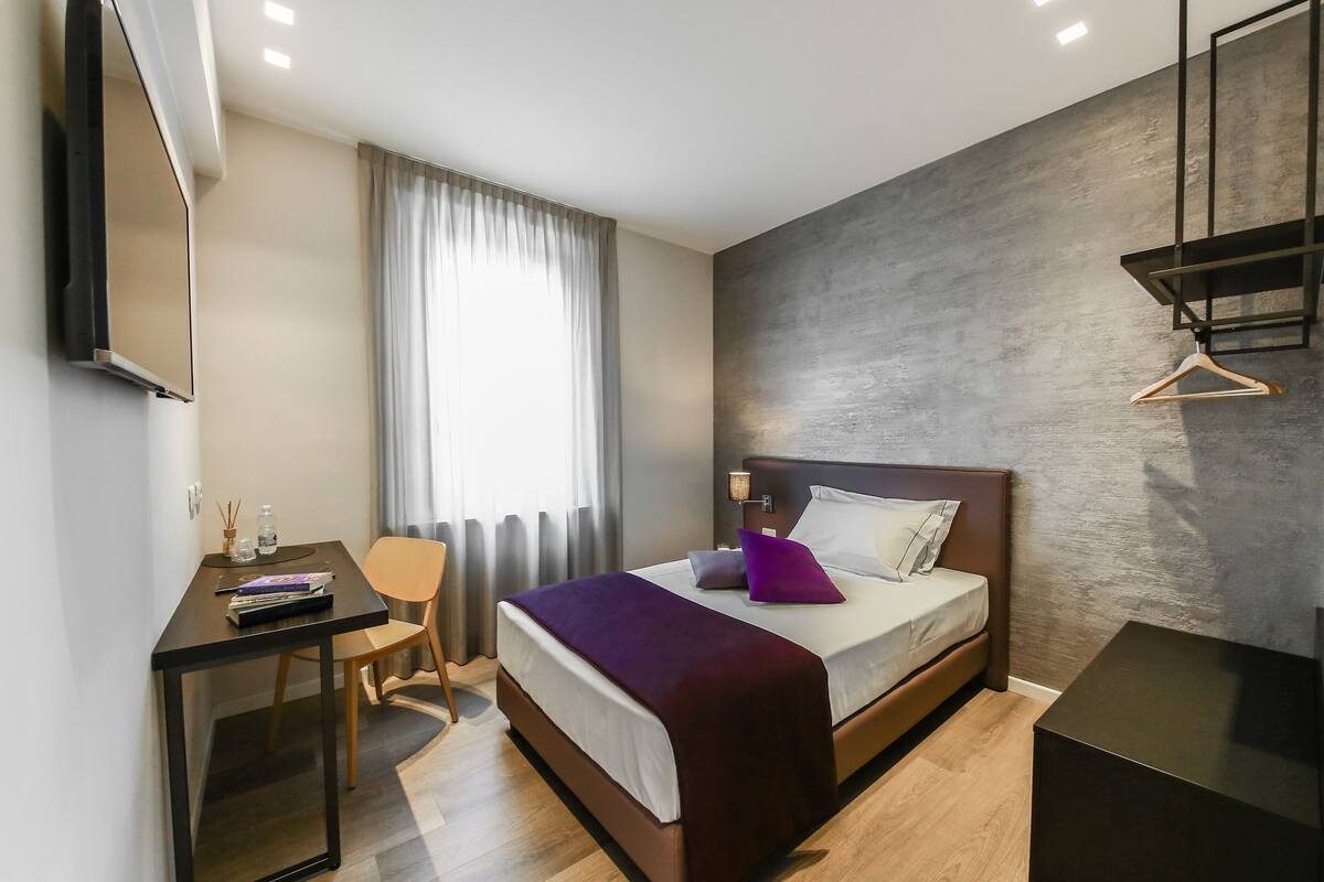 A modern hotel room with a bed, bedside table, desk, chair, and a TV mounted on the wall.