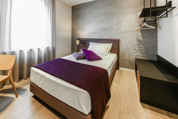 Quarto de hotel moderno com uma cama grande, uma mesinha de cabeceira, uma cadeira e um guarda-roupa.