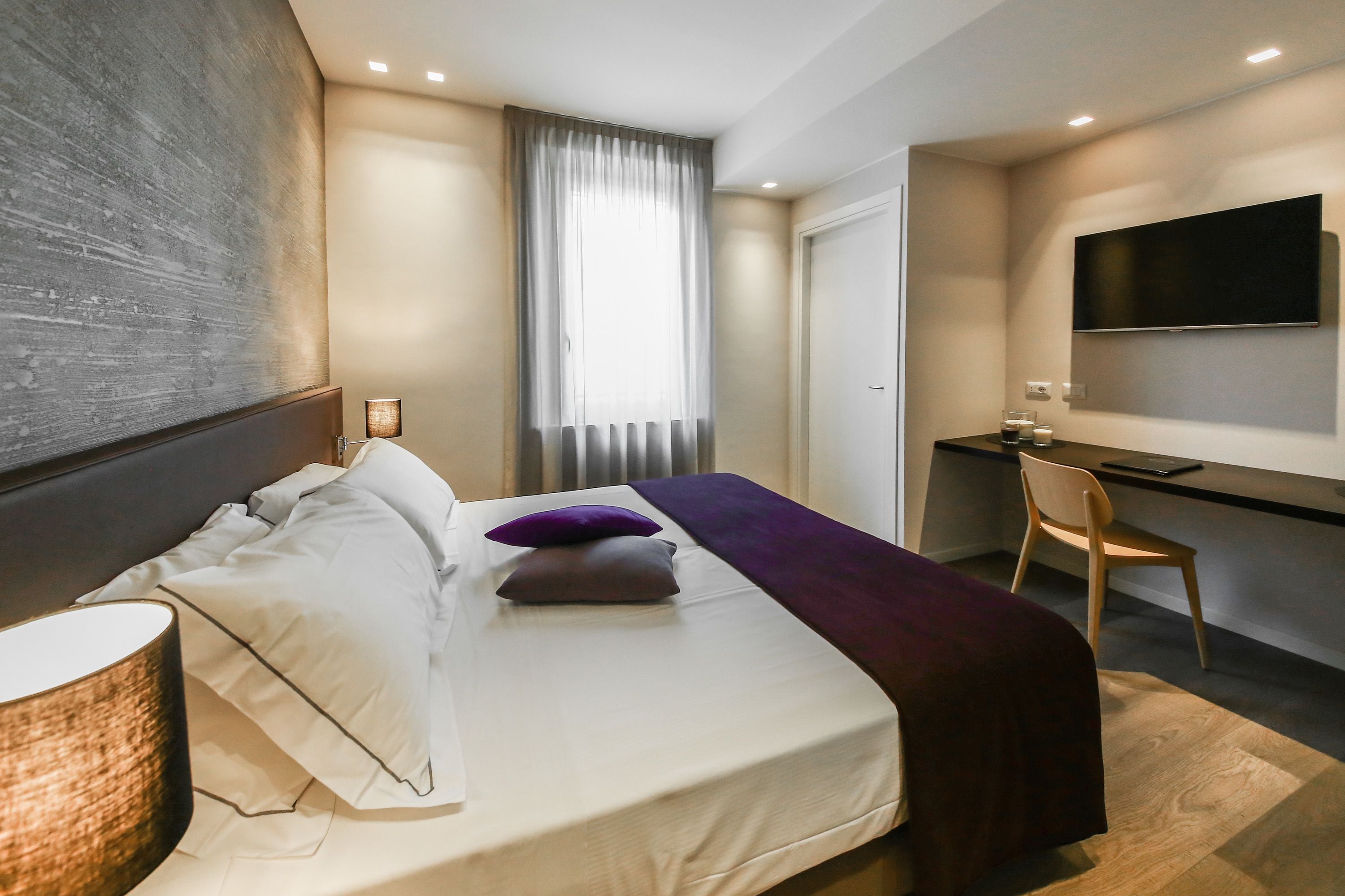 Una habitación de hotel moderna con cama, lámpara de noche, escritorio, silla y televisor montado en la pared.