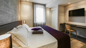 A modern hotel room with a bed, bedside lamp, desk, chair, and a TV mounted on the wall.