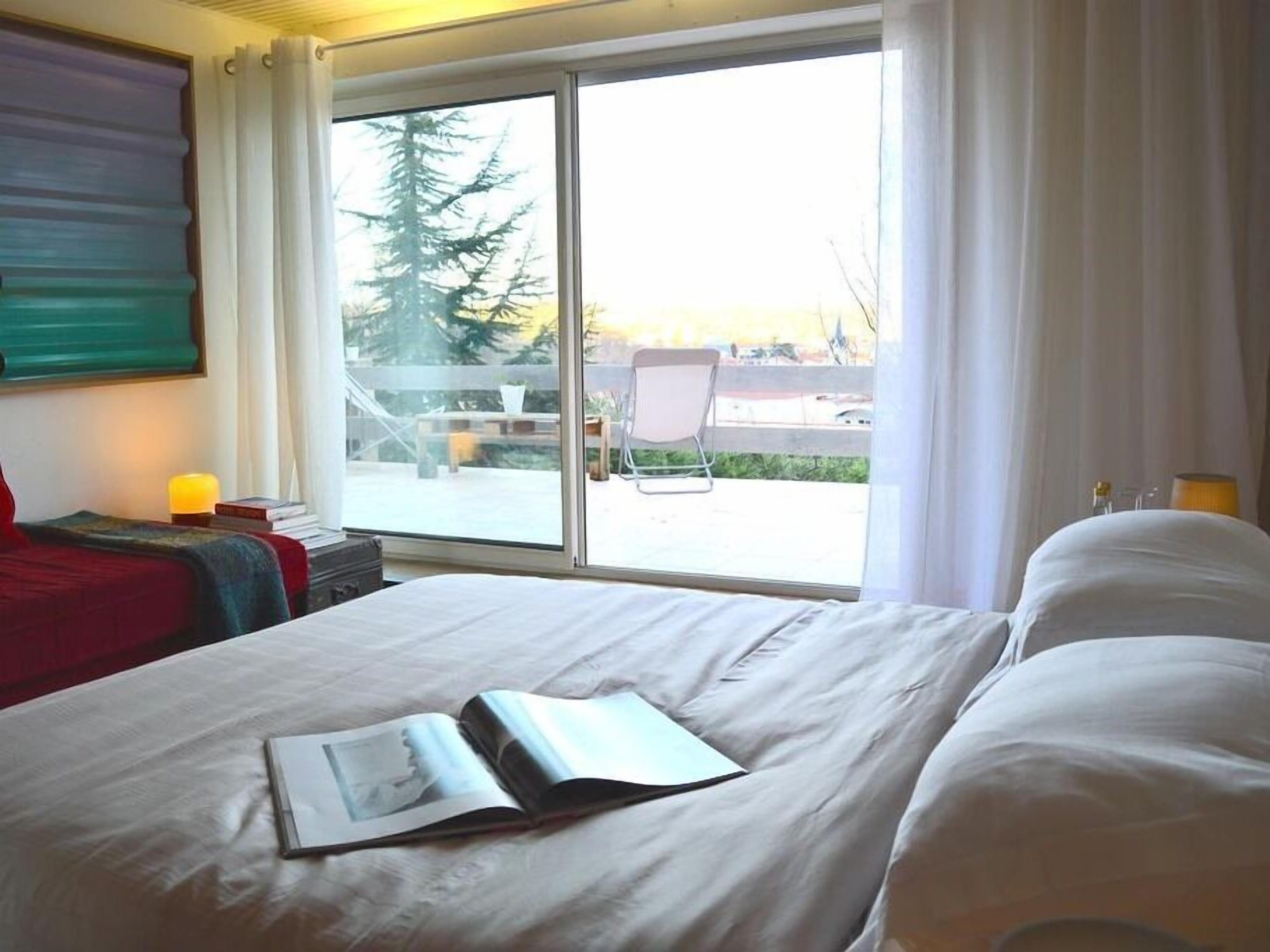 Habitación de hotel con un ventanal grande, una cama con ropa blanca, una silla y una mesita de noche con lámpara.