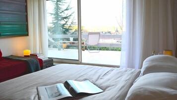 Habitación de hotel con un ventanal grande, una cama con ropa blanca, una silla y una mesita de noche con lámpara.