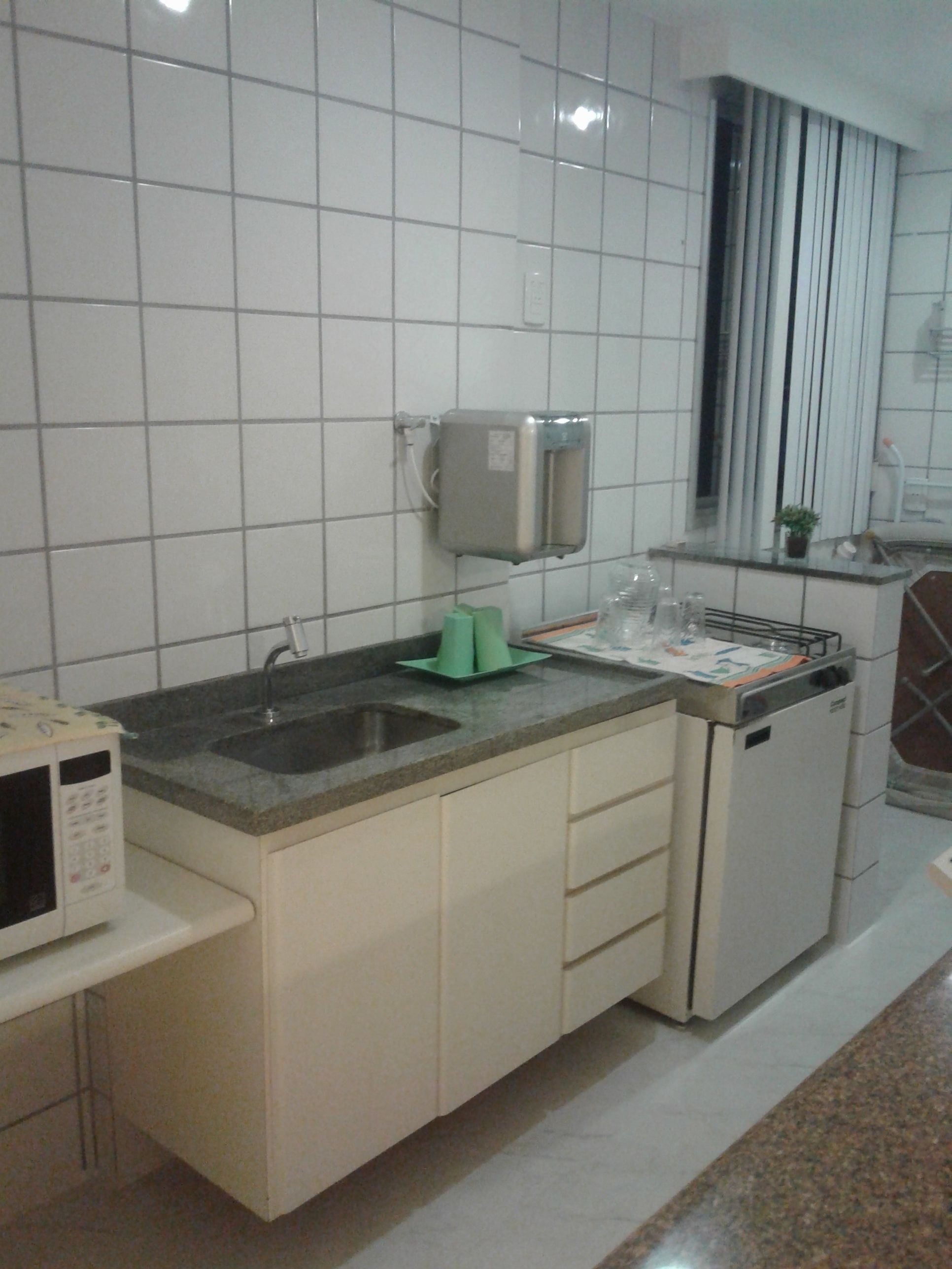Uma cozinha com armários brancos, bancada de granito, micro-ondas, pia e janela com persianas.