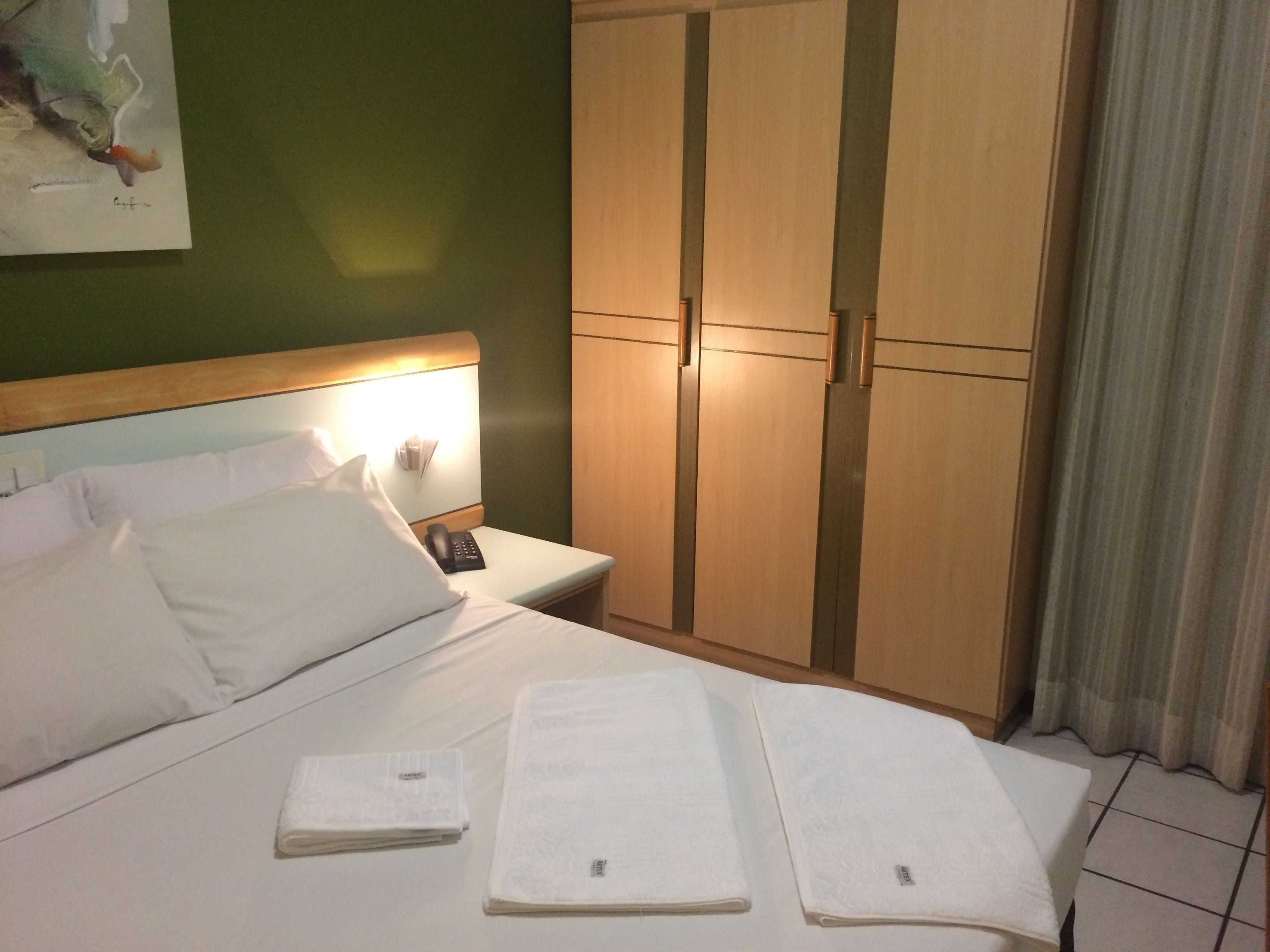 A hotel room with a bed, a nightstand with a lamp, a wardrobe, and a painting on the wall.