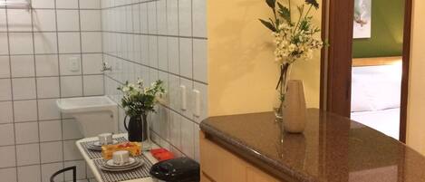 A compact kitchen with white tiled walls, a wooden countertop, a dining table with chairs, and a vase with flowers.