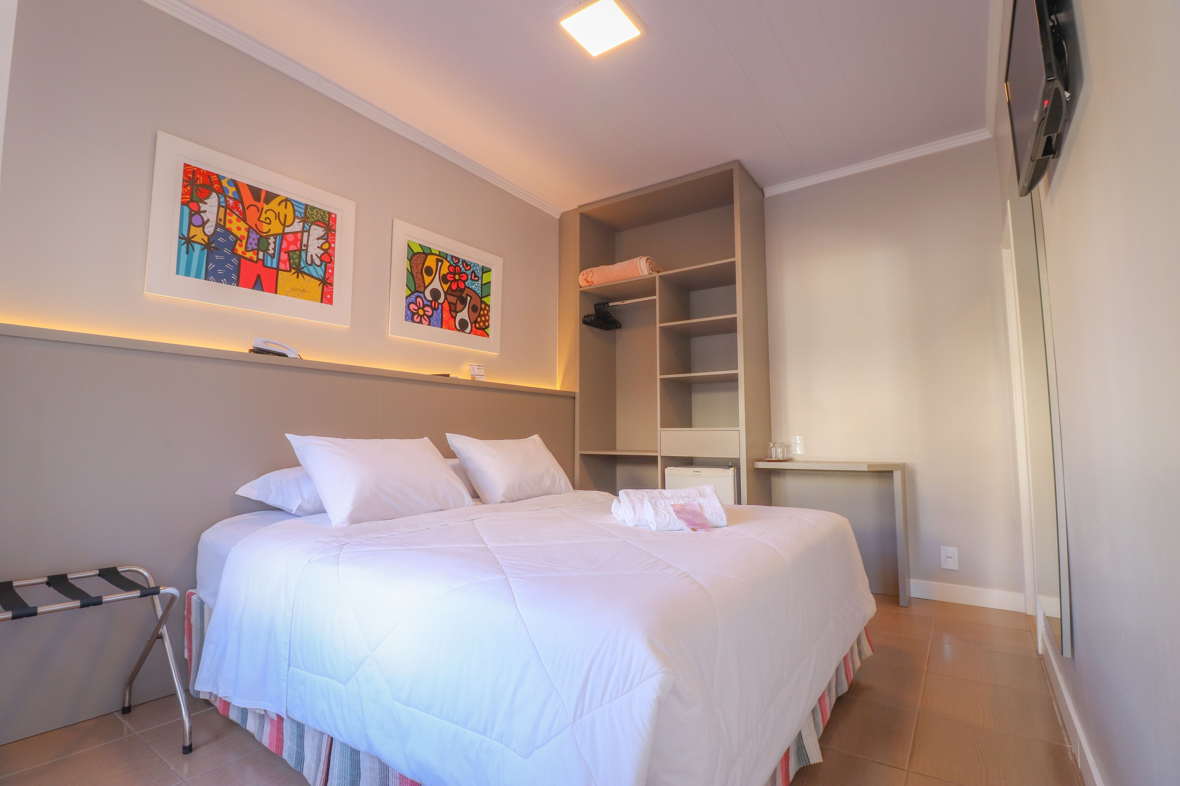 A neatly made bed with white linens, a small bedside table, and two colorful abstract paintings on the wall.
